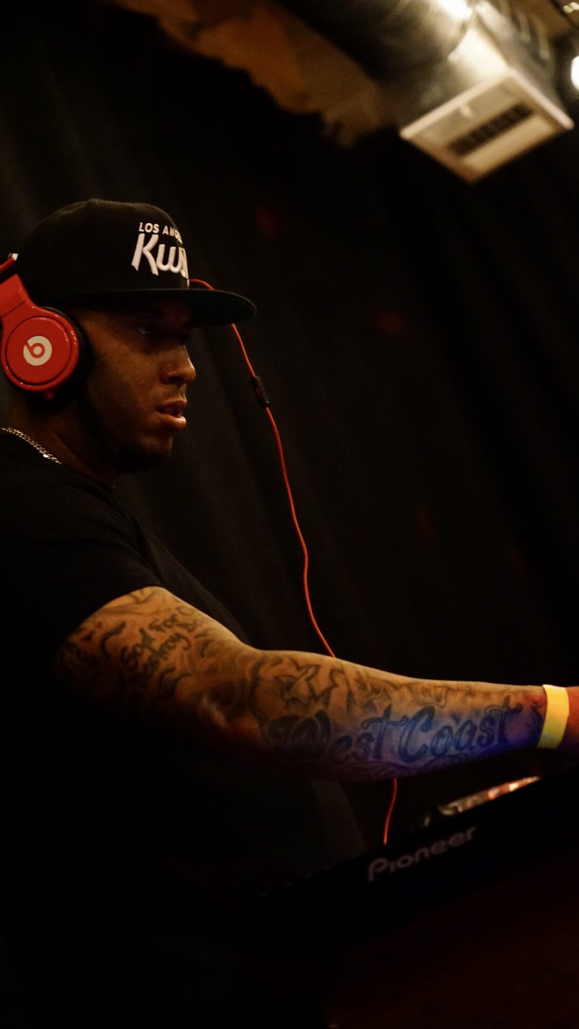 Person wearing a cap and red headphones, with tattoos on arm, in a dark setting.