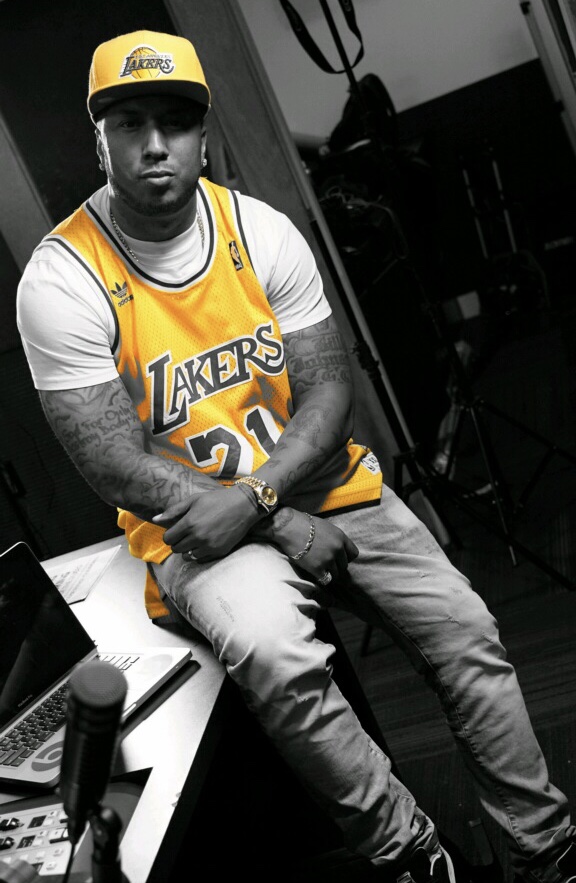 Young man wearing a yellow Lakers jersey and cap, sitting with arms crossed, in a room with equipment and a laptop.