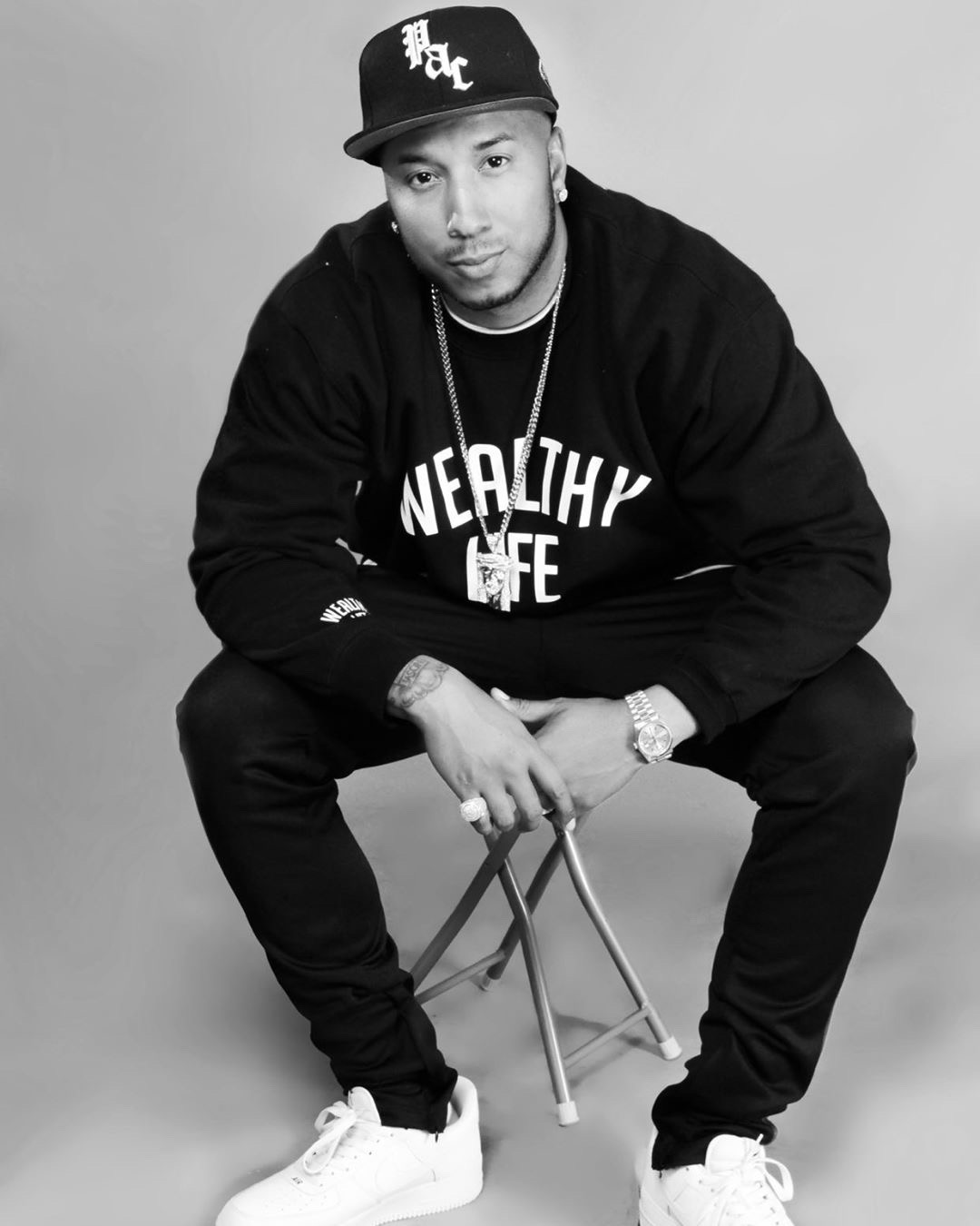 Man sitting on a small stool, wearing a cap, hoodie, and sneakers, looking at the camera in a studio setting.