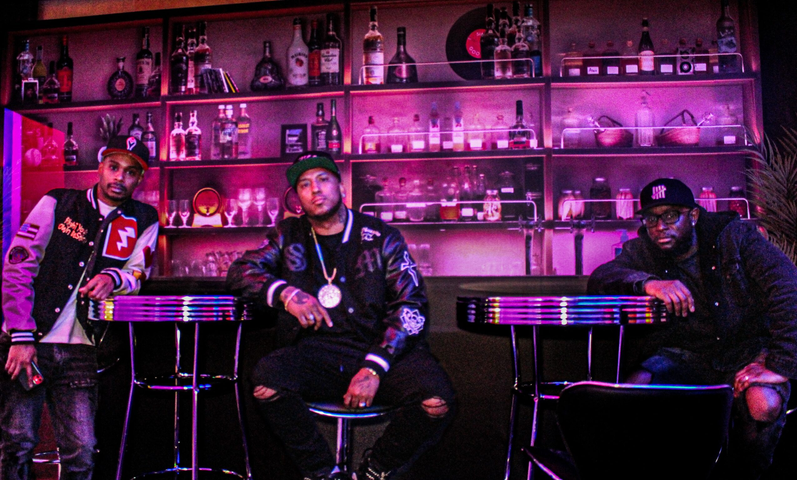 Three men sit at a bar with shelves of bottles illuminated in pink and purple lighting behind them.
