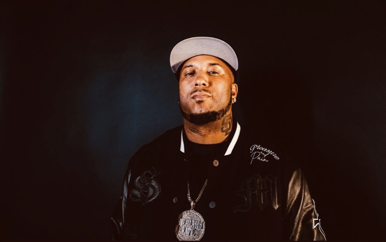 Person wearing a gray cap, black jacket, and a large pendant necklace, standing against a dark background.