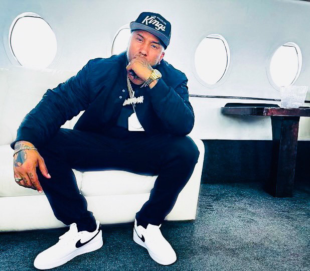 Person sitting on a white seat inside an airplane, wearing a cap, dark jacket, white sneakers, and jewelry, with airplane windows in background.