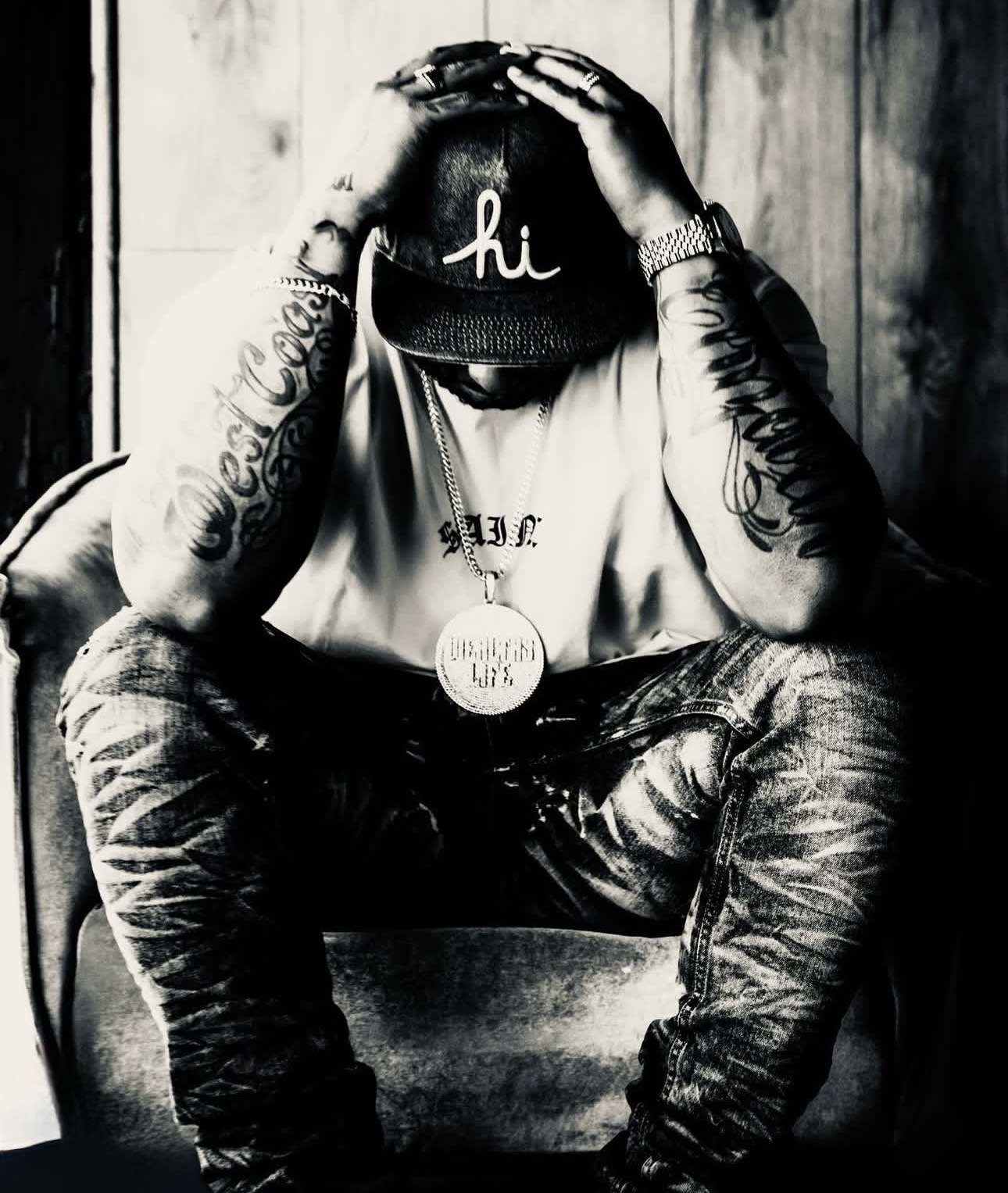 Person sitting with head down, hands on head, wearing a cap, tattoos visible on arms, in black and white.