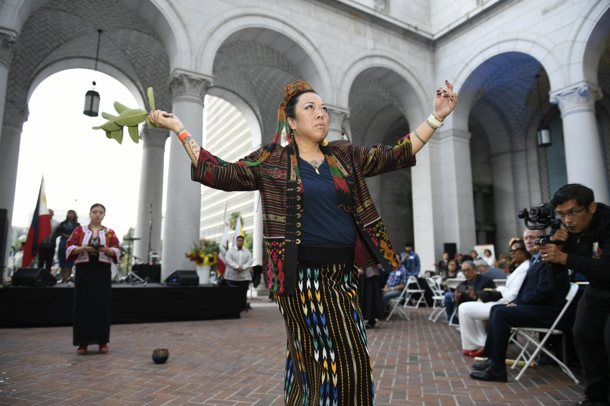 Woman with arms raised holding a plant, performing in a courtyard with audience and photographers present.