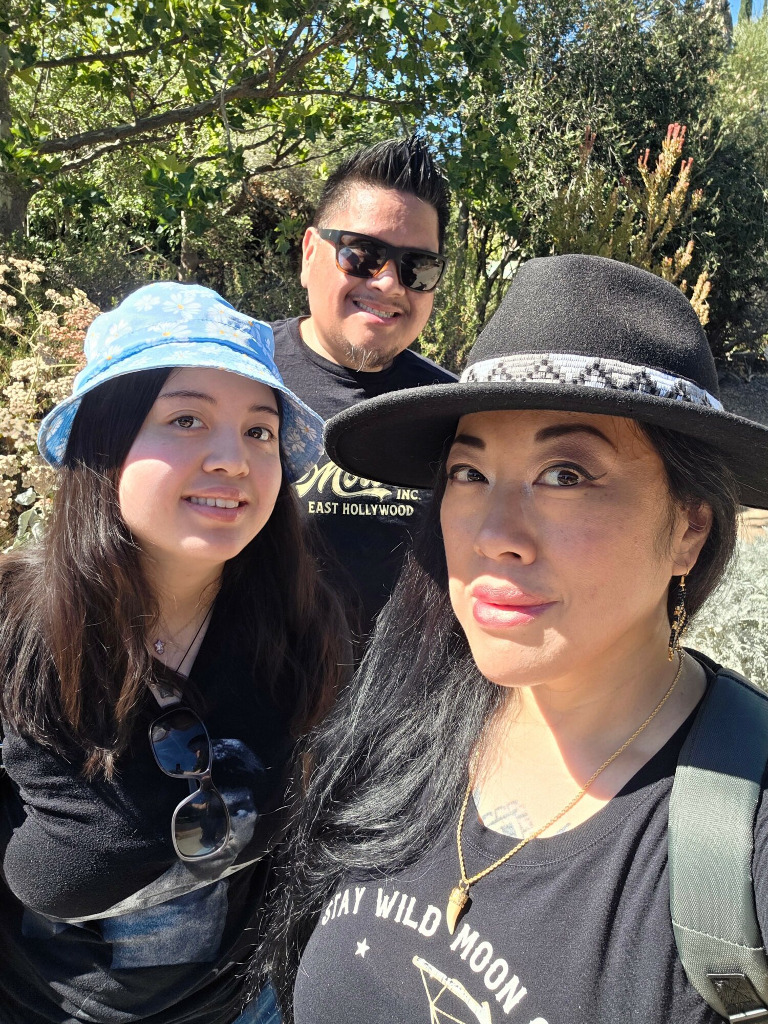 Three people outdoors, two women and one man, smiling, with trees and sunlight in the background.