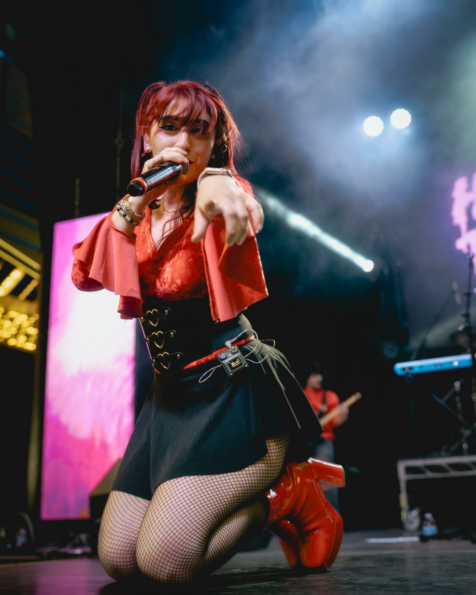 Performer with red hair singing into microphone on stage, wearing red top, black skirt, fishnet stockings, and red boots, with stage lights.