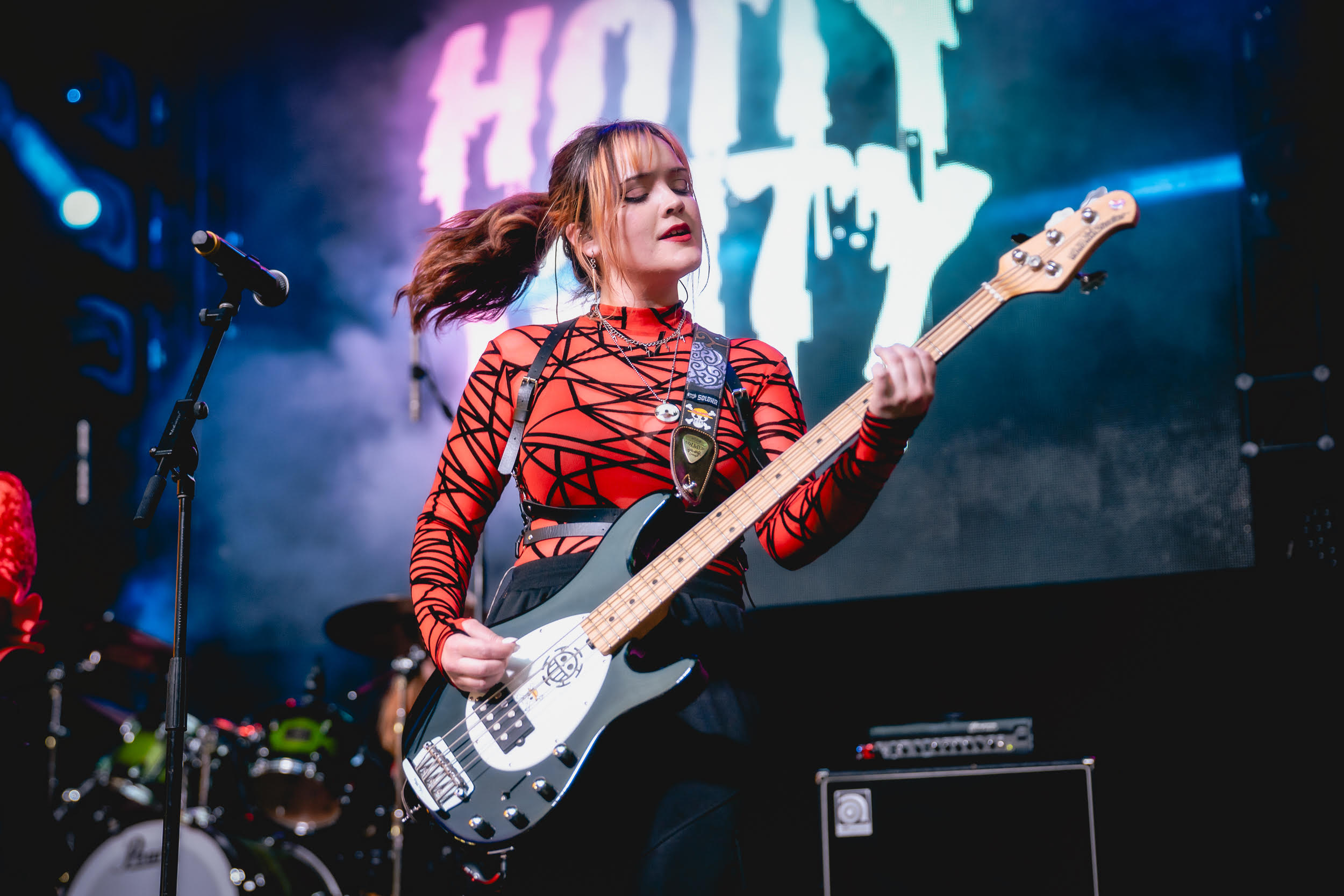 Female musician playing an electric guitar on stage with a large screen behind her displaying text.