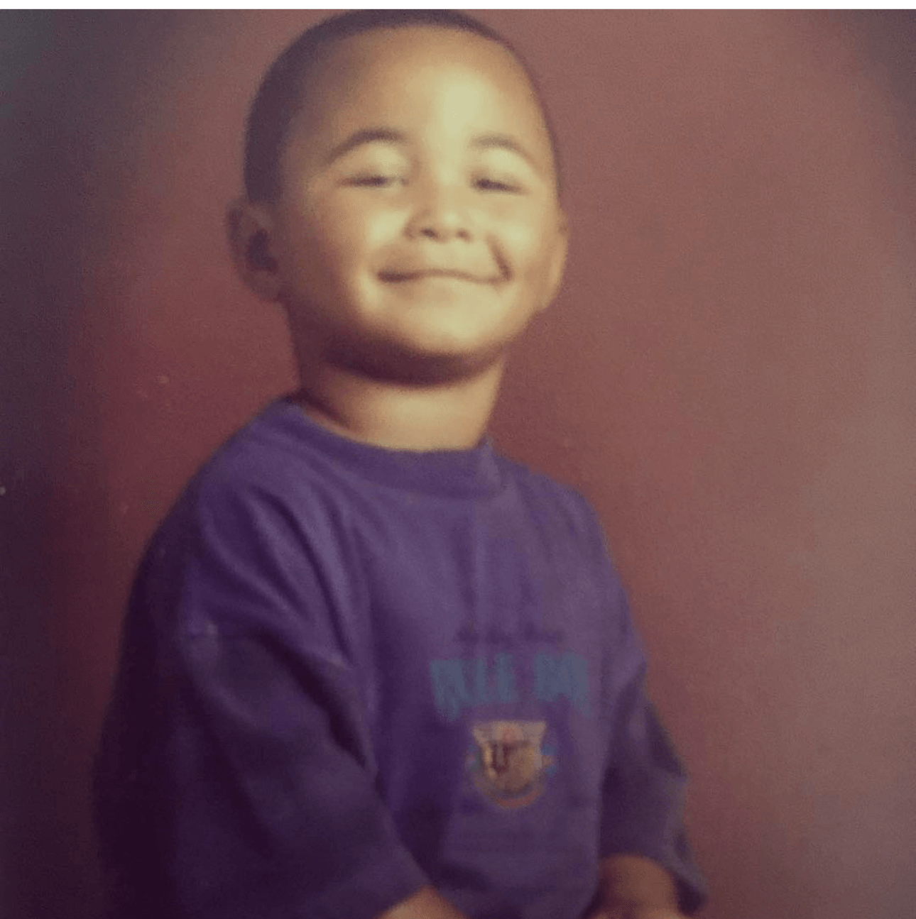 Child with short hair smiling, wearing a purple shirt, against a plain background.