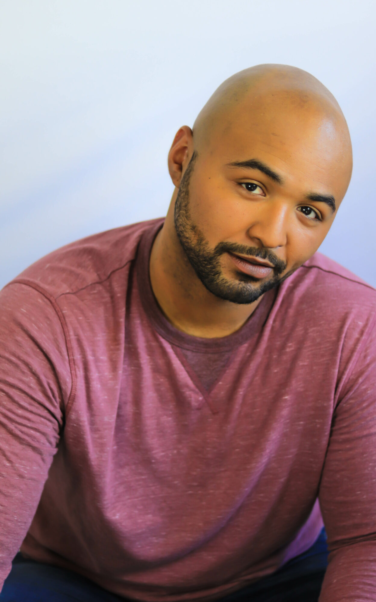 A man with a shaved head and beard wearing a maroon shirt, looking at the camera with a neutral expression.