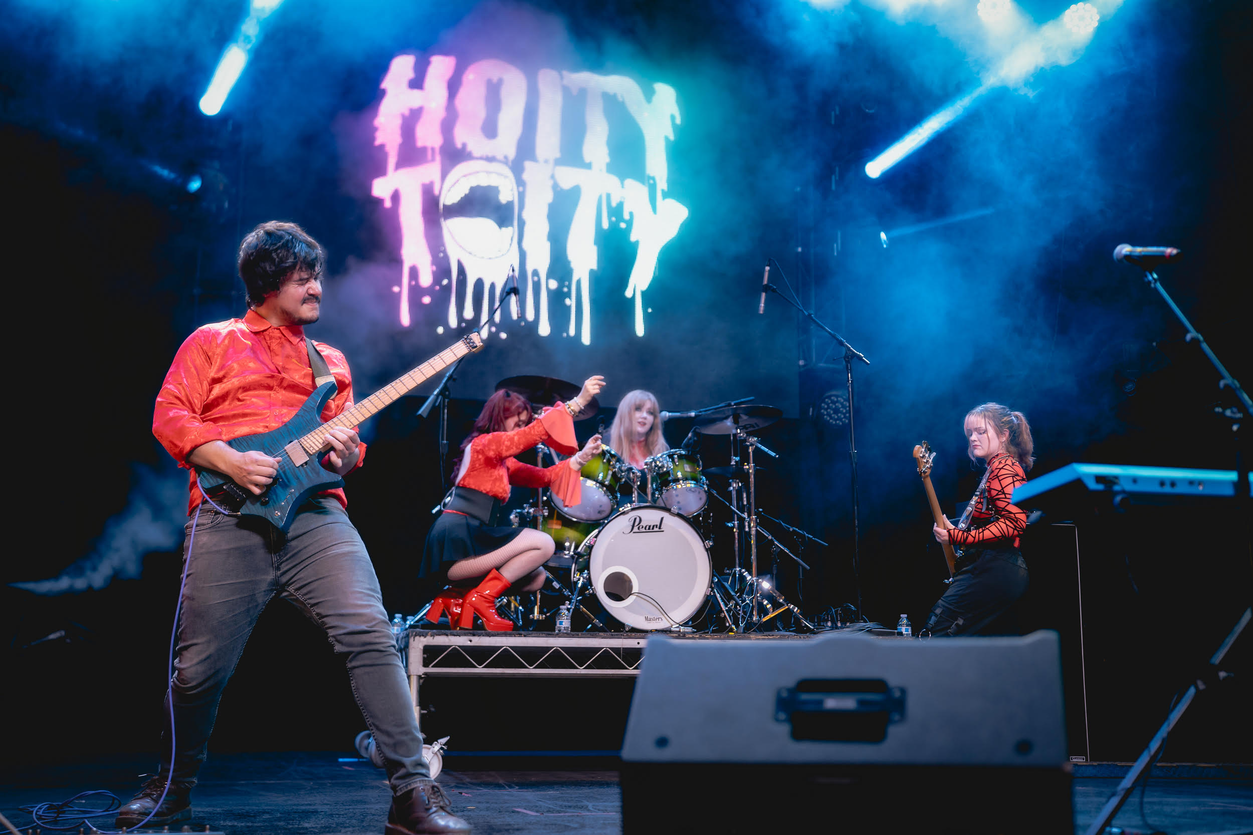 Four musicians perform on stage with a colorful sign reading 'HOLLY CITY' in the background.