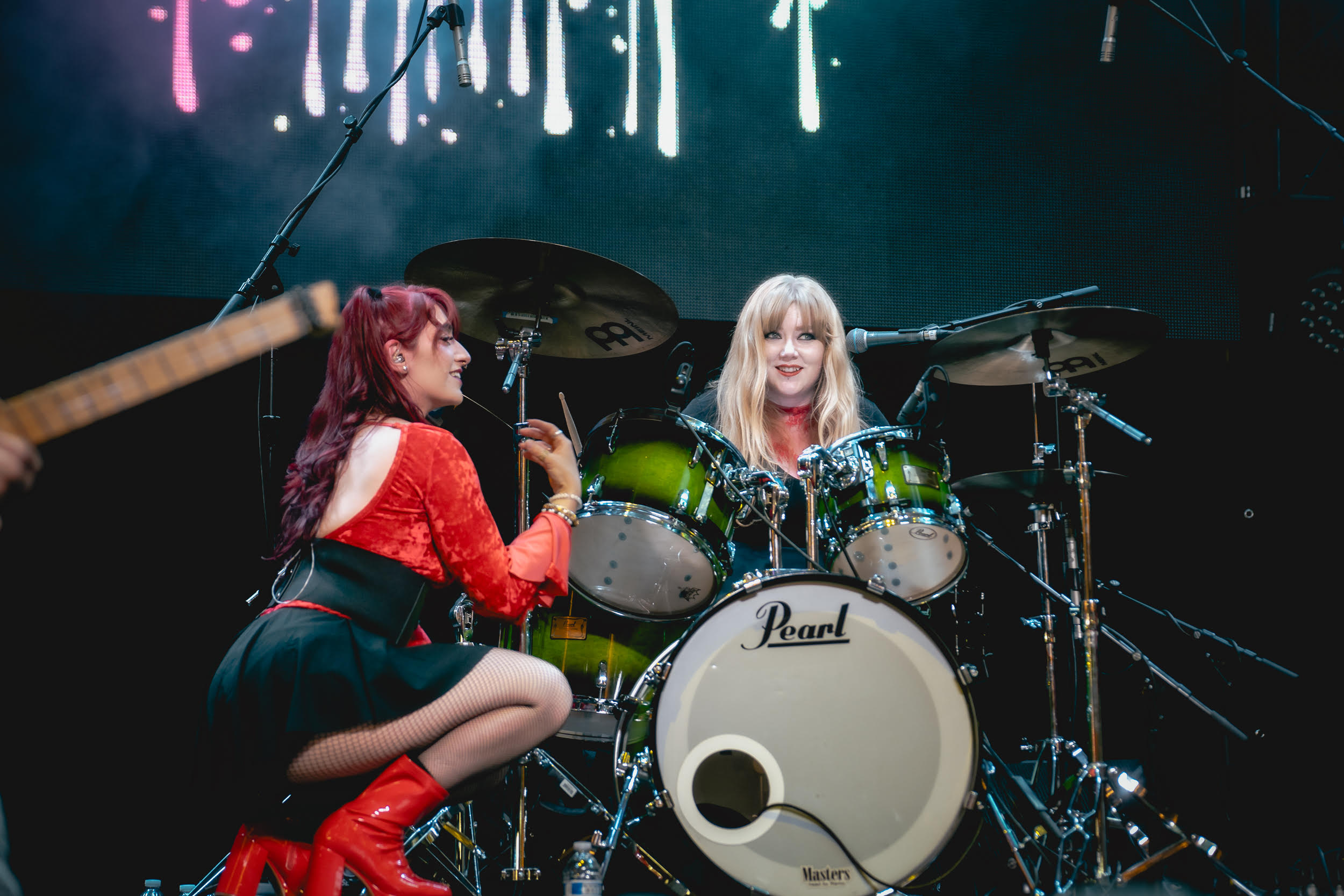 Two women performing on stage with a drum set, one singing into a microphone, stage lights in background.