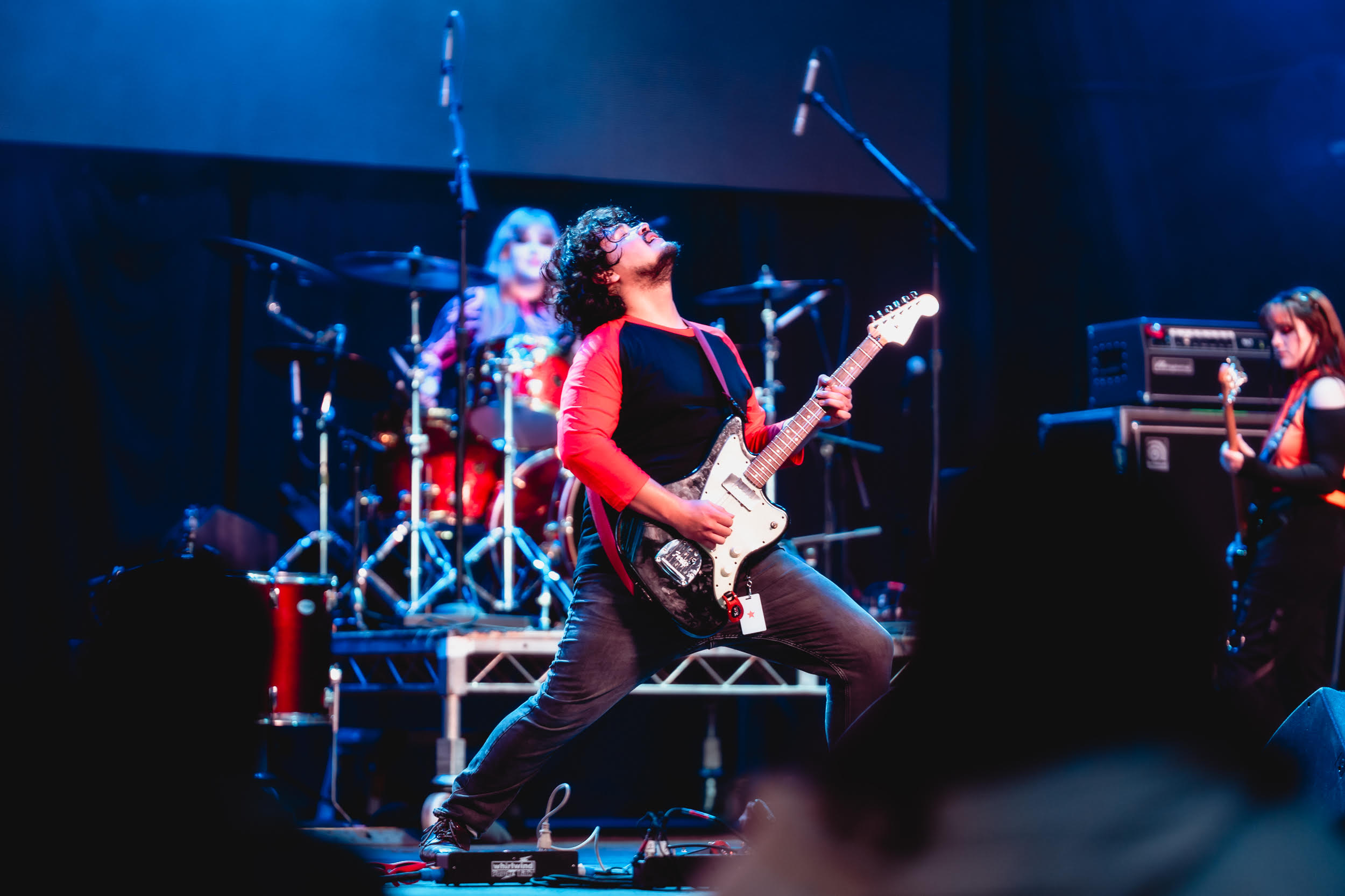 Three women perform on stage with musical instruments, including a guitarist in the center, a drummer in the background, and a keyboardist on the right.