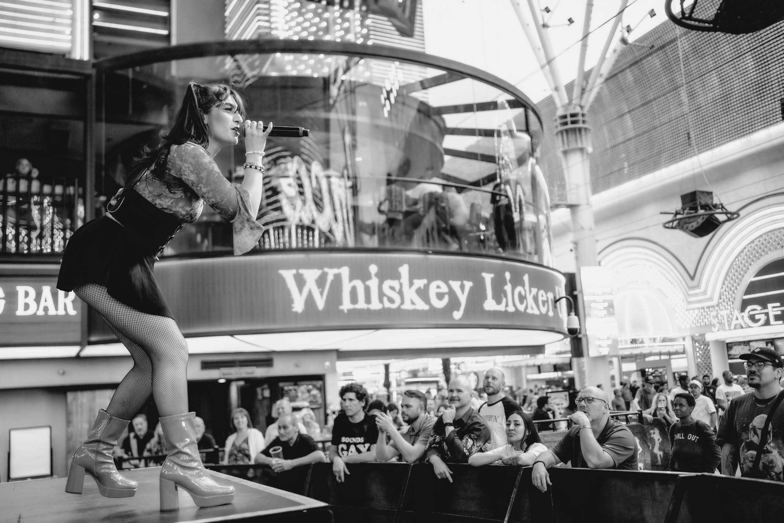 A woman in a short dress and boots leans on a bar counter with a sign reading 'Whiskey Licker' inside a busy indoor venue.