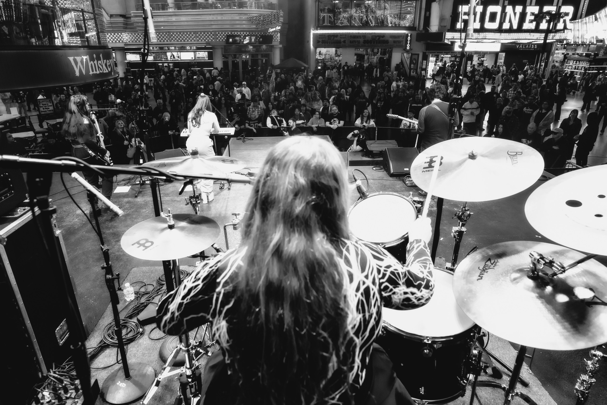Person with long hair playing drums on stage in front of a crowd in a shopping mall.