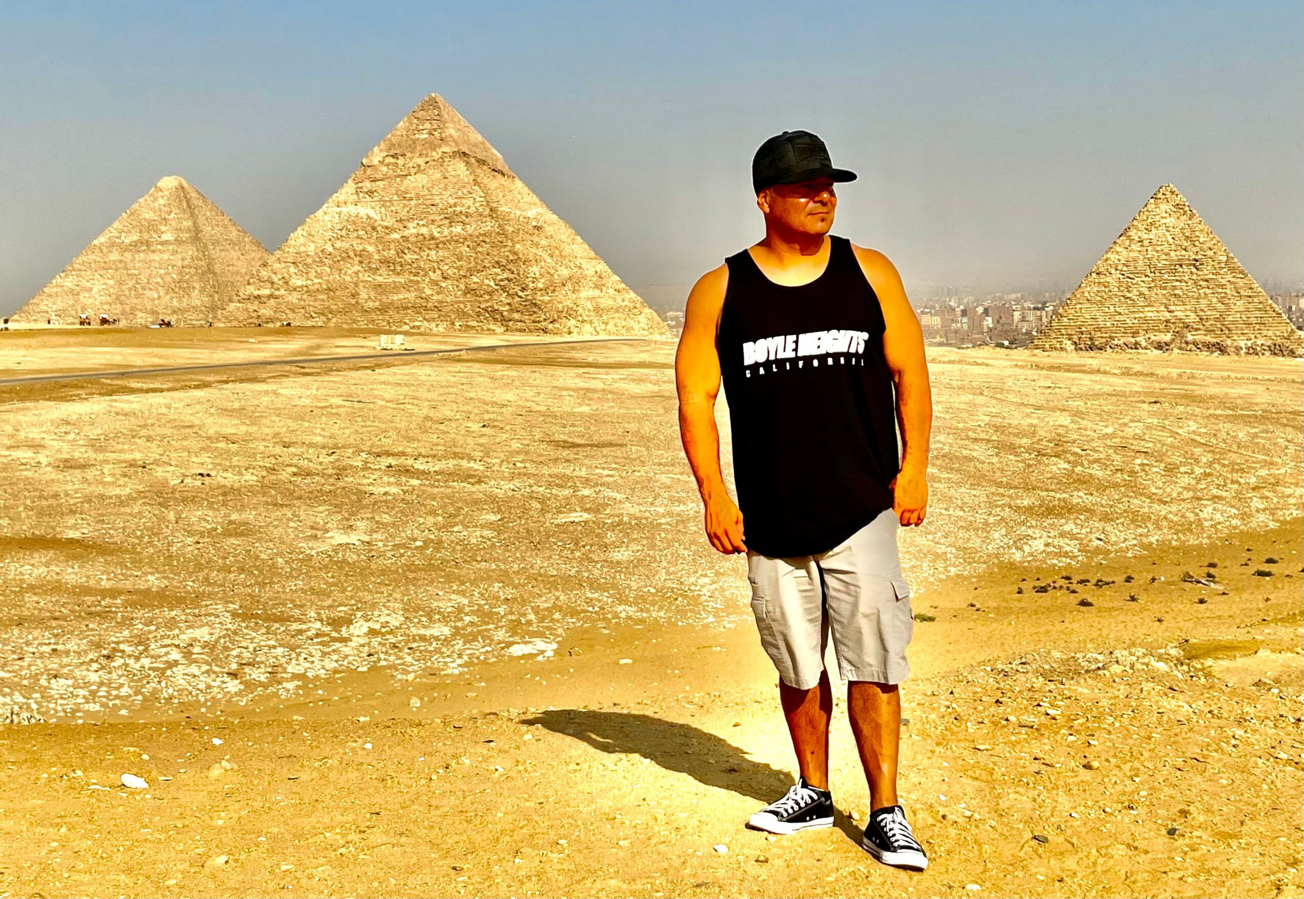 Person standing in desert with pyramids in background, wearing a black cap, sleeveless shirt, and shorts, casting a shadow.