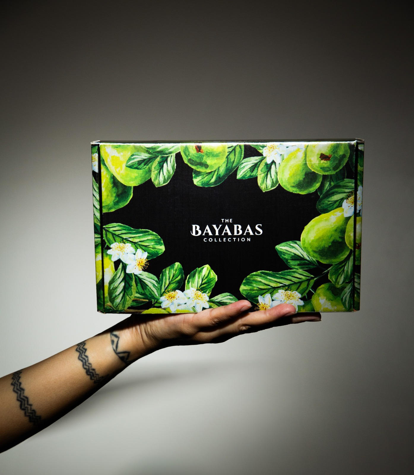 A hand with a tattoo holds a box with green leaves and white flowers, labeled 'The Bayabas Collection'.