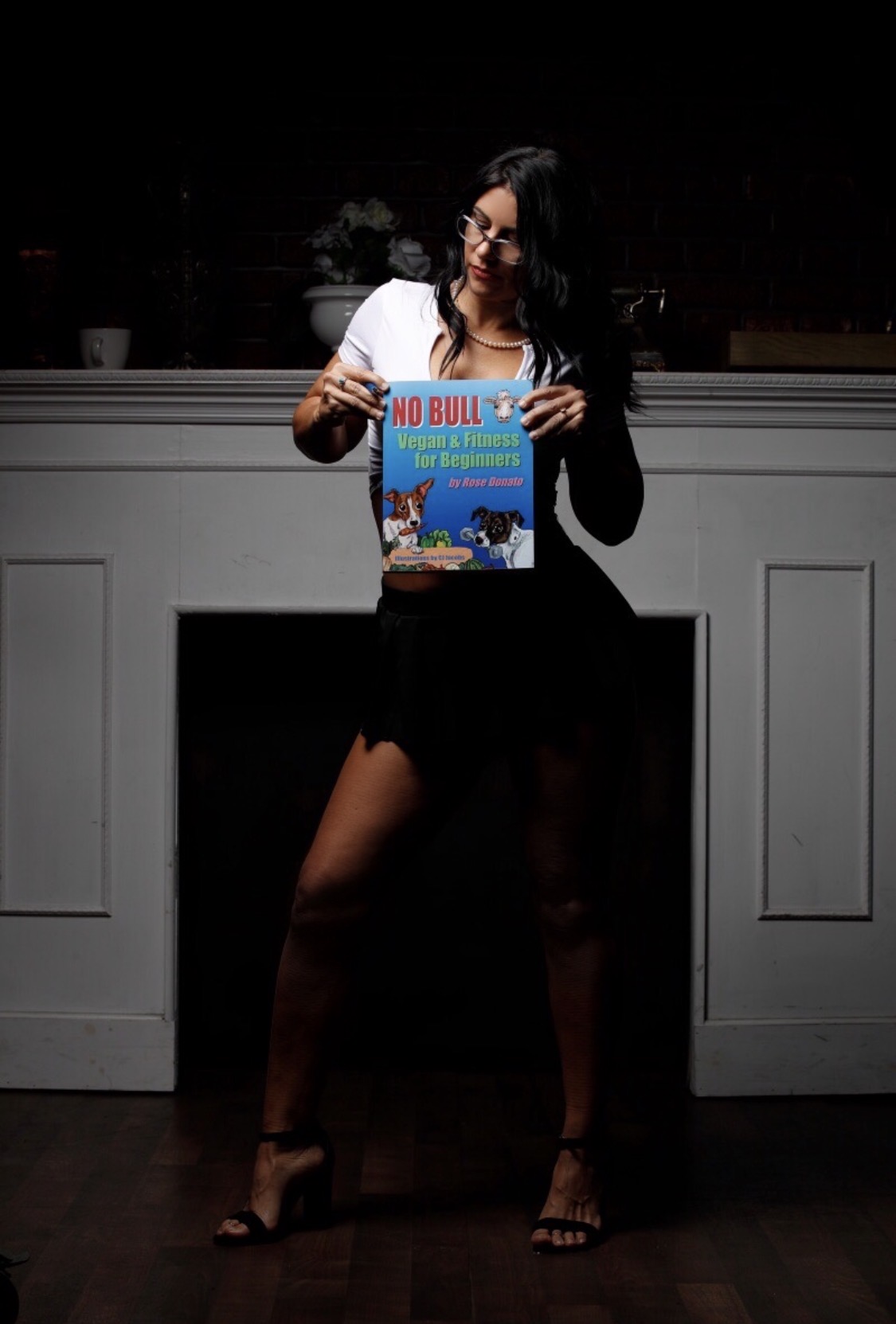 Woman holding a No Bull magazine, standing indoors with a dark background, smiling, wearing a white top and black skirt.