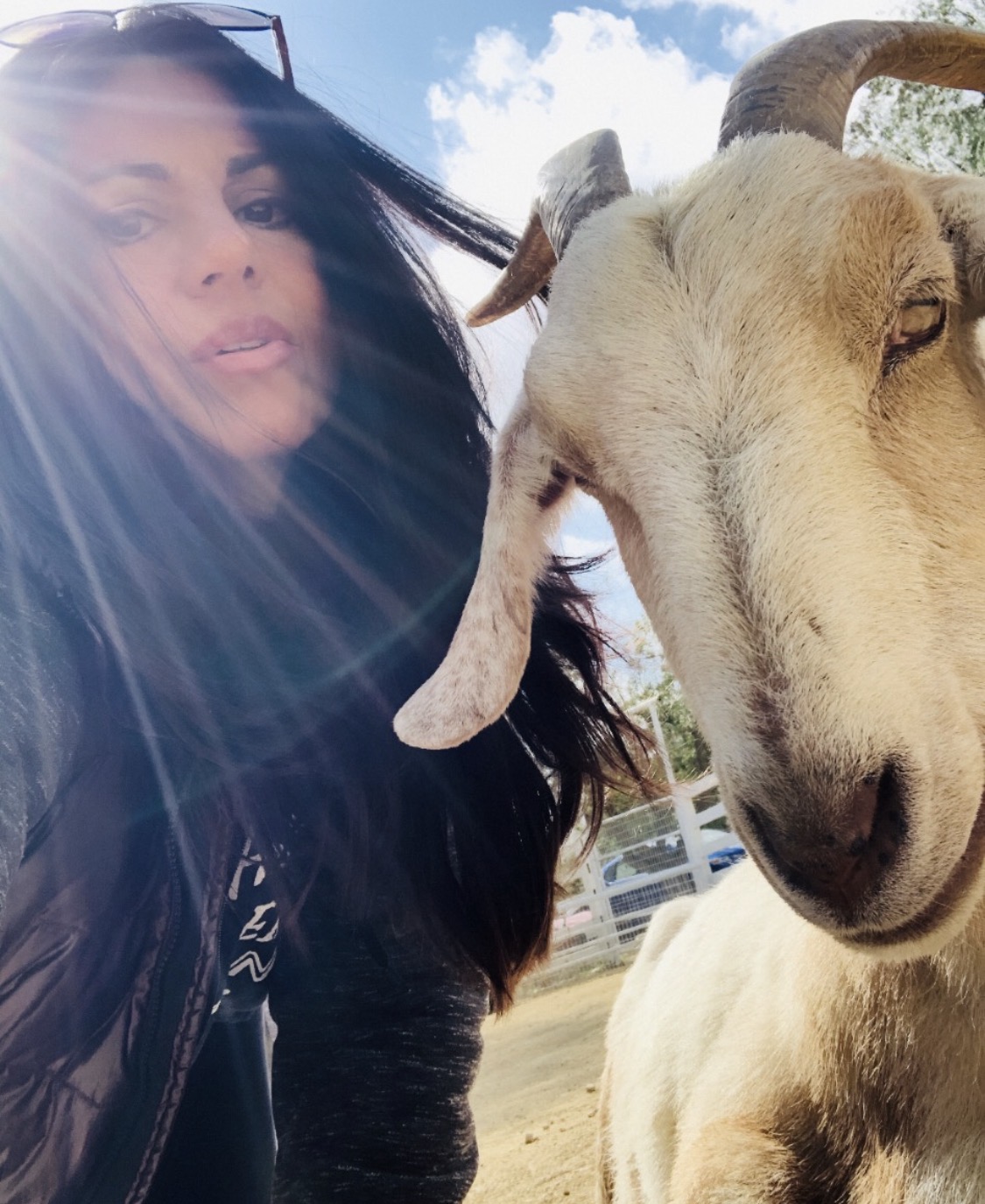 Person with long dark hair and sunglasses taking a selfie with a goat outdoors, blue sky and clouds in background.