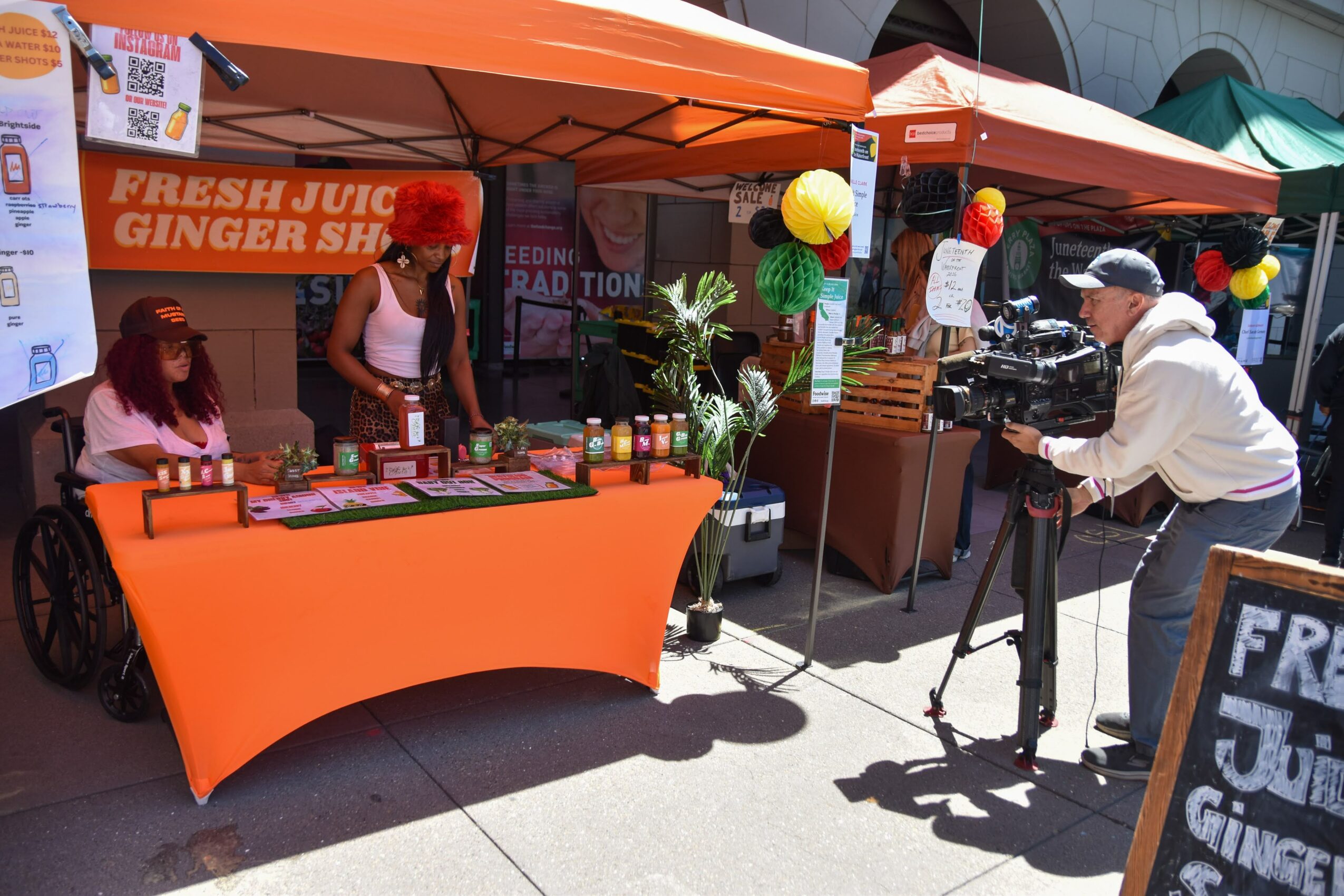 Person filming a woman at a juice stand with orange tablecloth and banner, outdoor market setting, bright sunlight, colorful decorations.