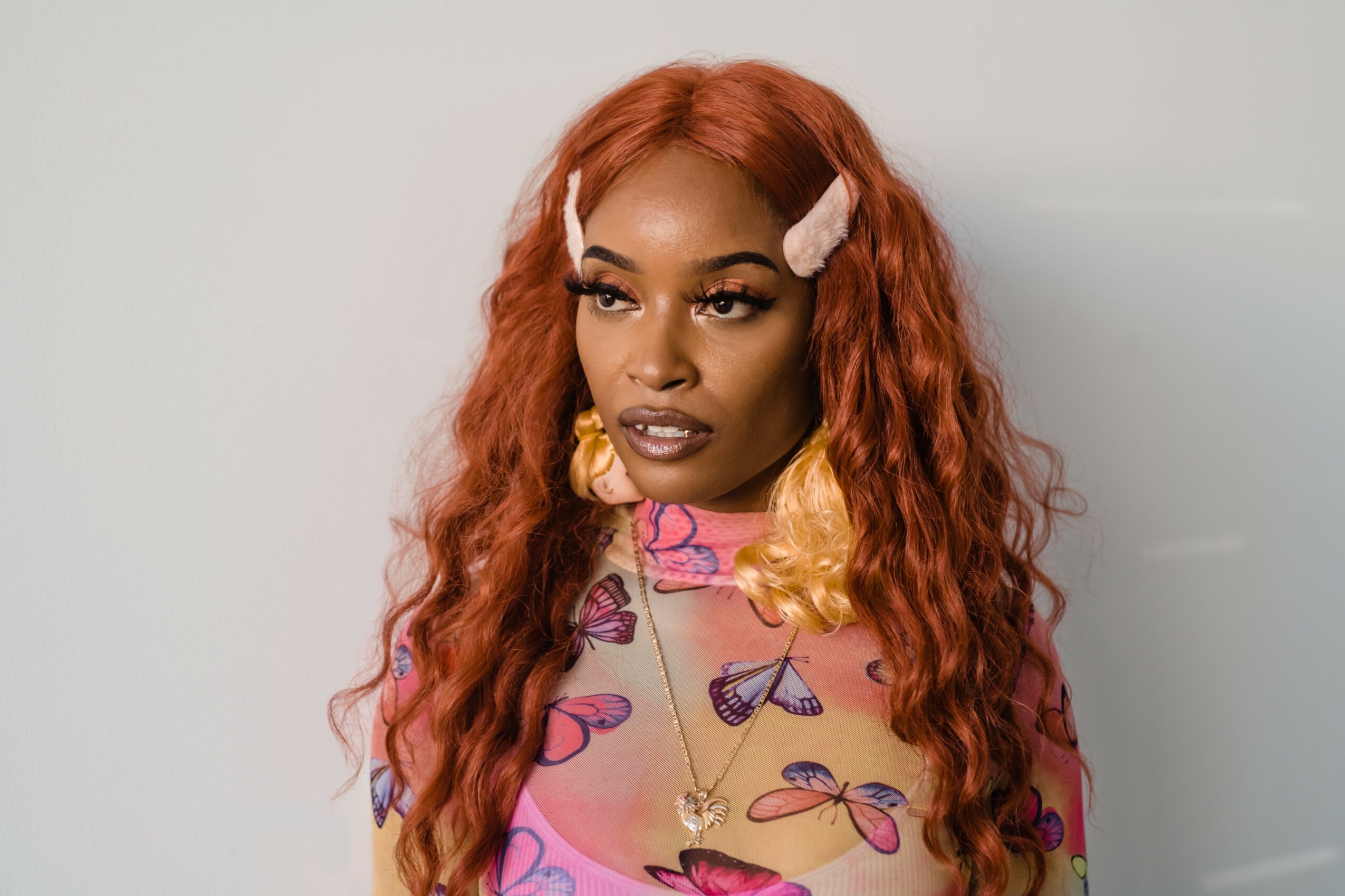 Woman with long, wavy red hair, wearing pink hair clips, colorful butterfly-patterned top, and large earrings, looking to the side.