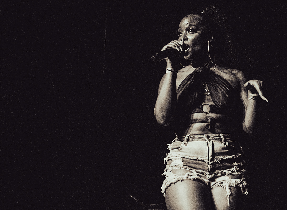 Young woman singing into a microphone on stage, wearing a crop top and ripped shorts, with a dark background.
