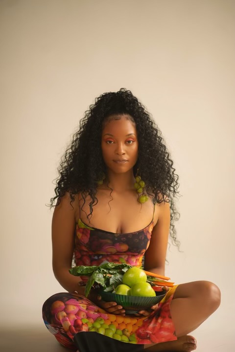 Young woman with curly hair sitting cross-legged holding a bowl of fruit, wearing a colorful dress, against a plain background.