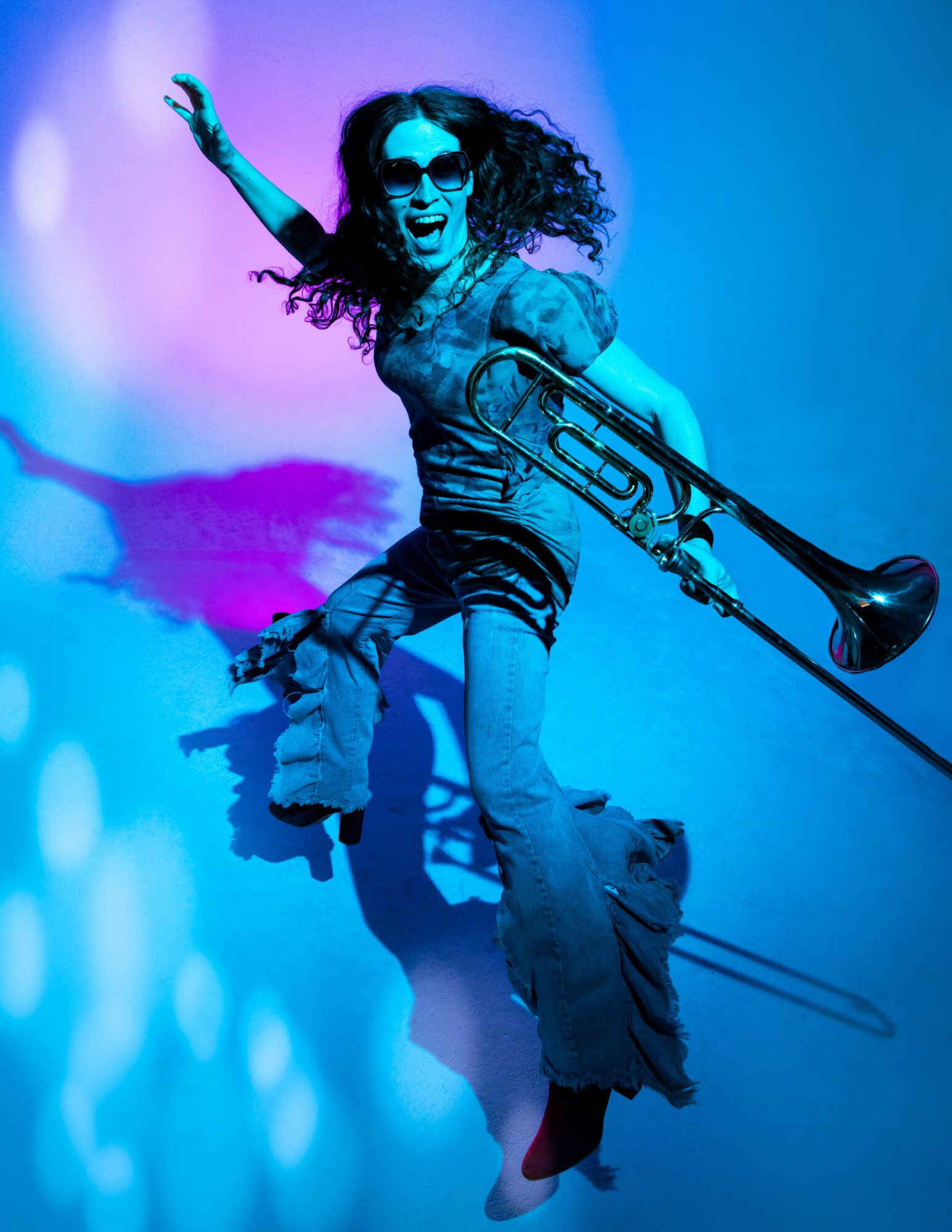 Person with curly hair wearing sunglasses, holding a trombone, jumping with a blue background.
