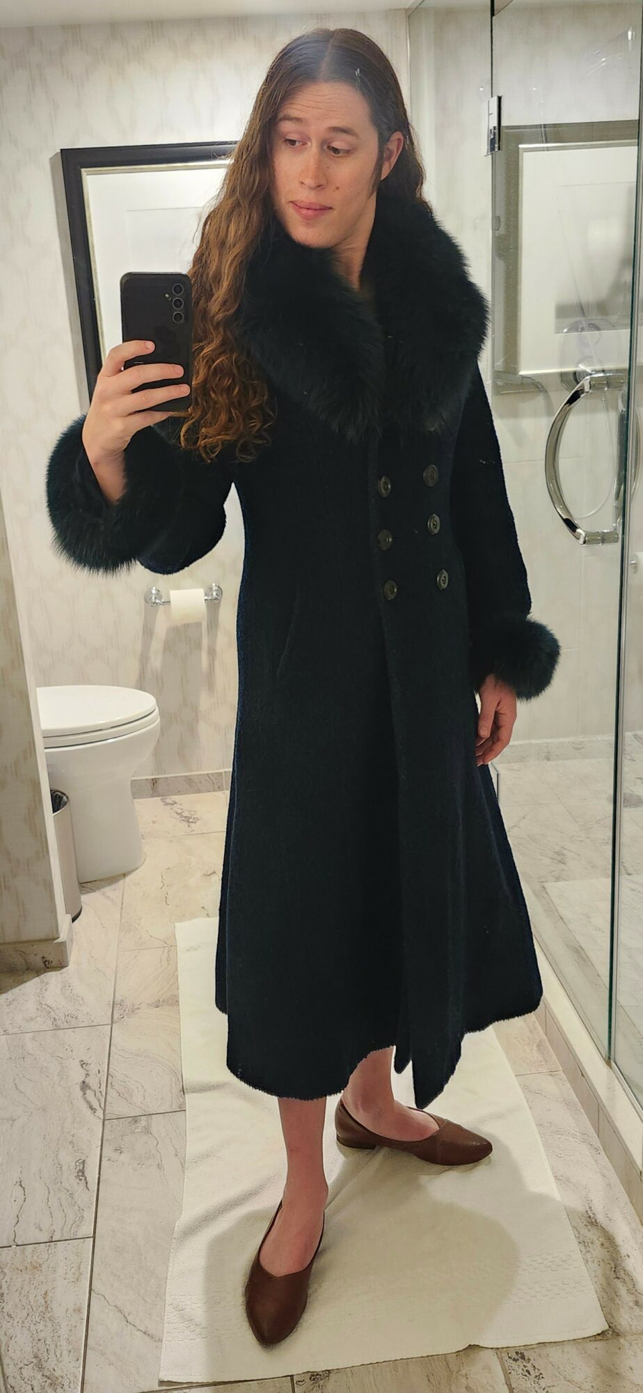 Woman taking mirror selfie in bathroom, wearing a long dark coat with fur collar, and brown shoes.