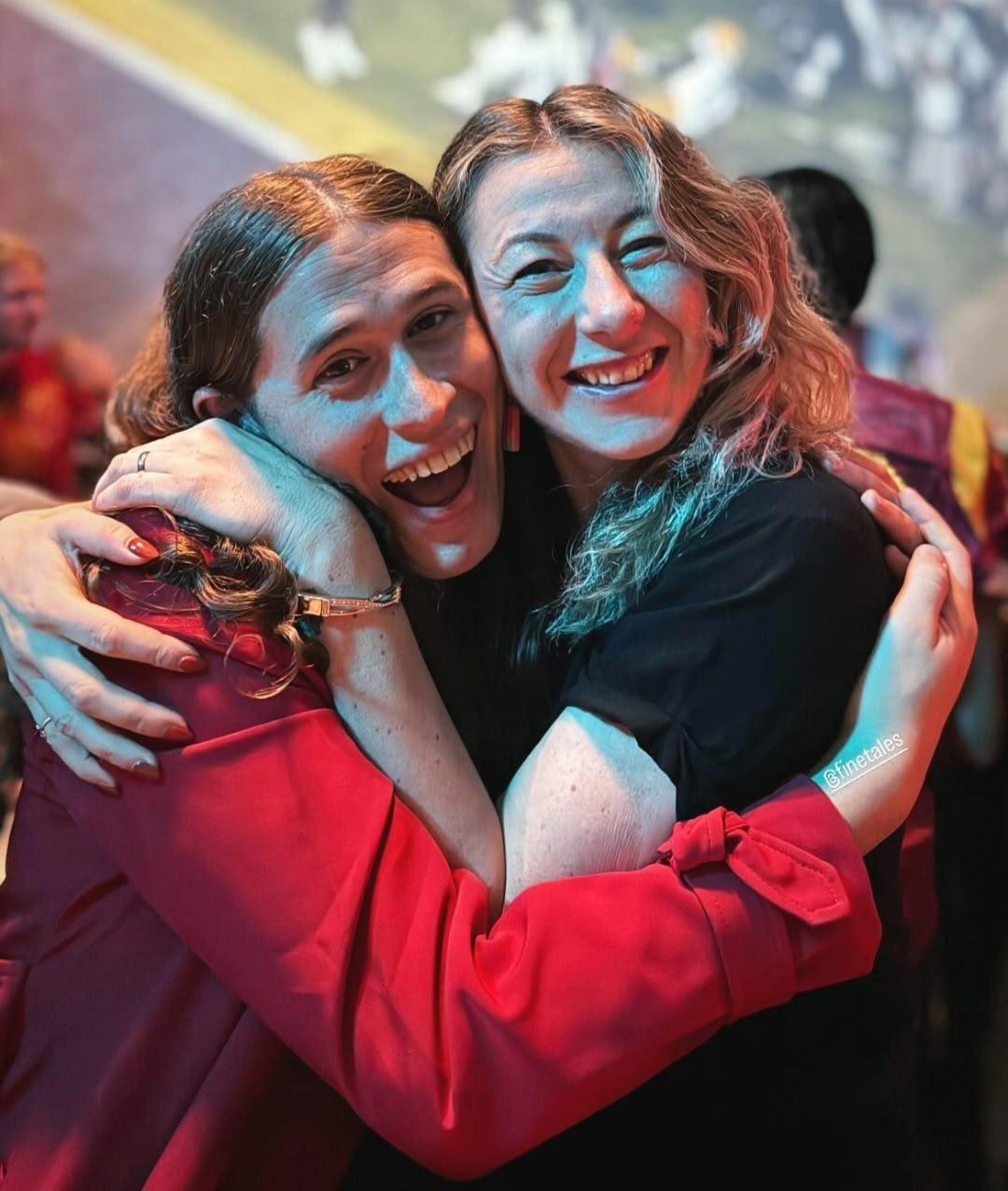 Two women hugging and smiling at a celebration or party, with colorful background lighting.