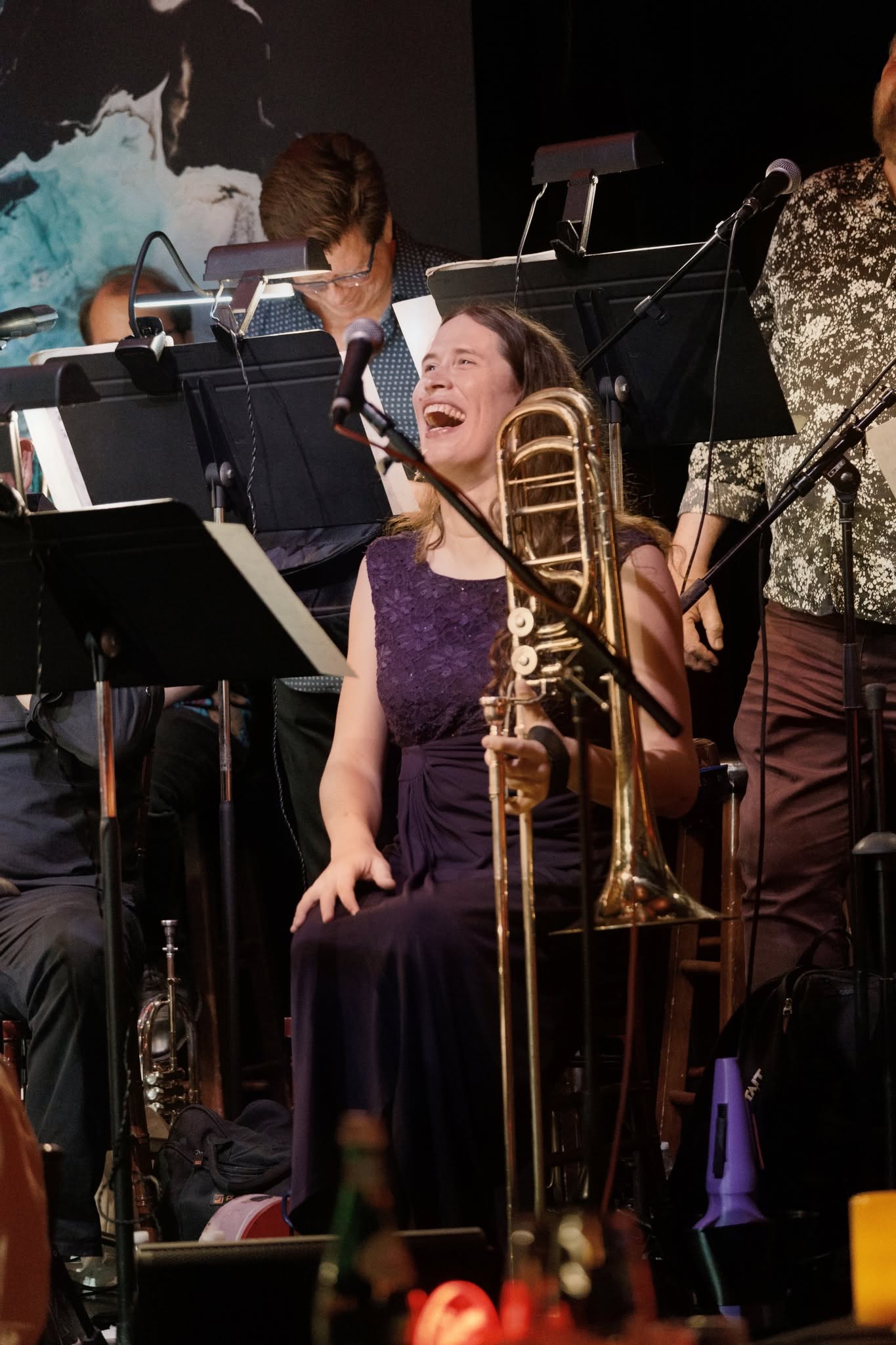Woman playing a trombone, smiling, surrounded by musicians with music stands, in a concert setting.