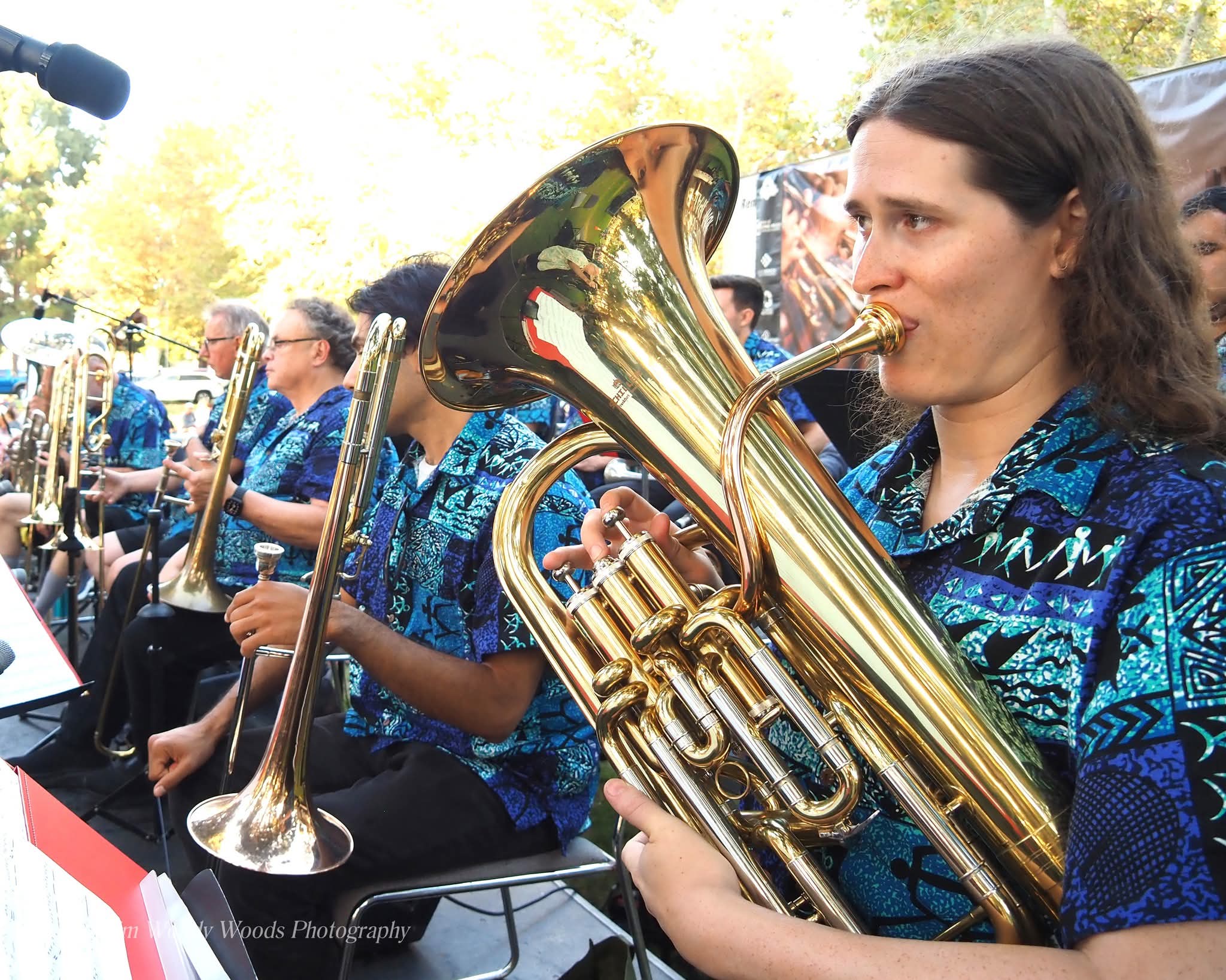 Woman playing a brass instrument outdoors with other musicians, trees in background, all wearing blue patterned shirts.