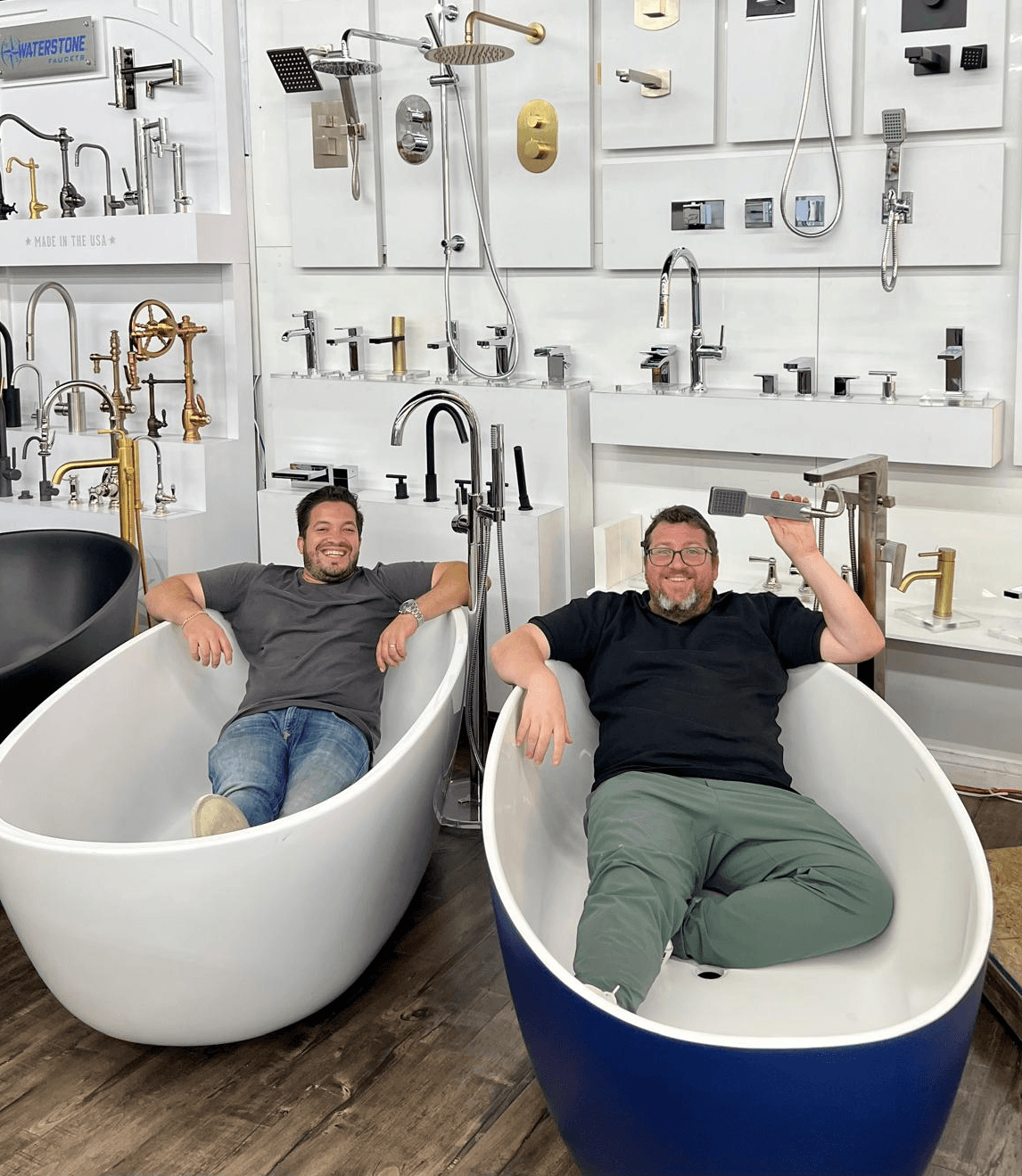 Two men relax in modern bathtubs in a showroom with various faucets and fixtures displayed on the wall behind them.