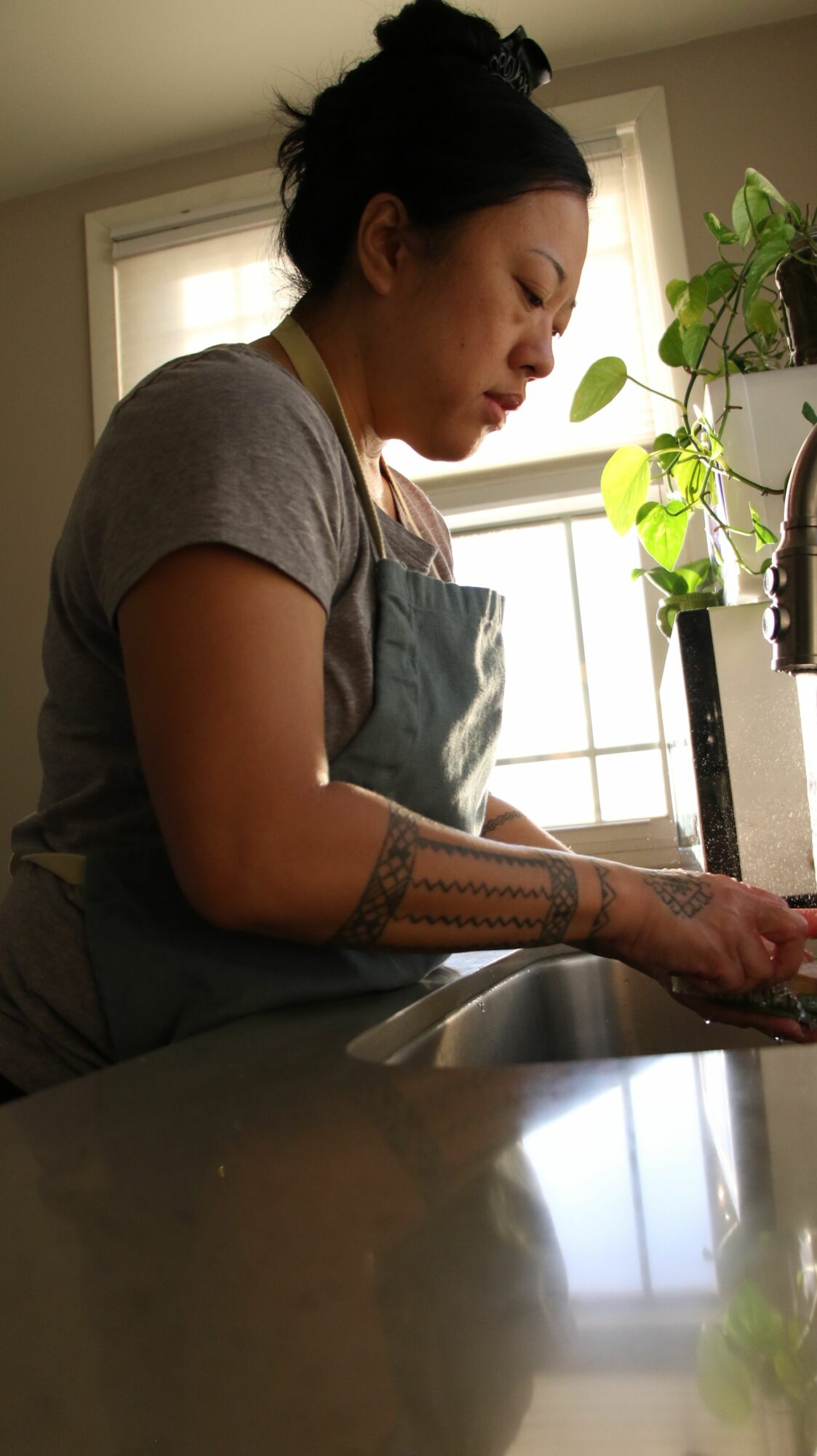 Person with tattoos leaning over a table near a window with plants, in a room with natural light.