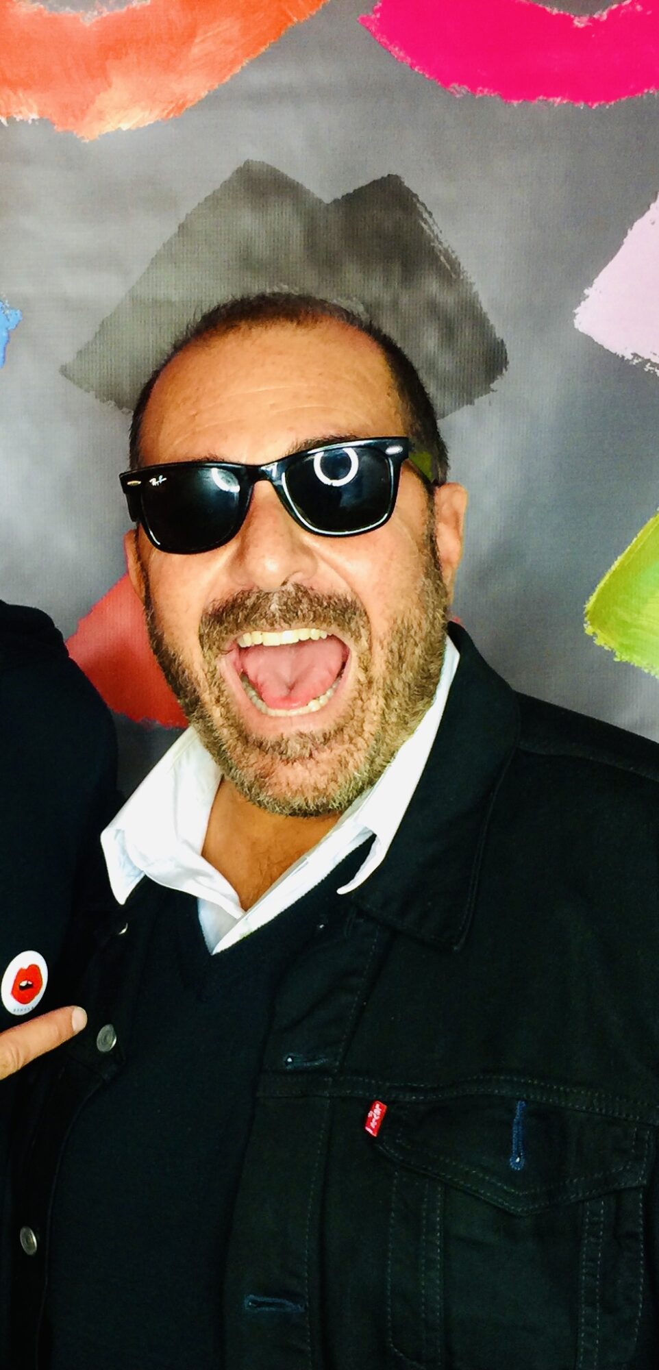 Man with sunglasses smiling, background with colorful abstract shapes, wearing a dark jacket and white shirt.