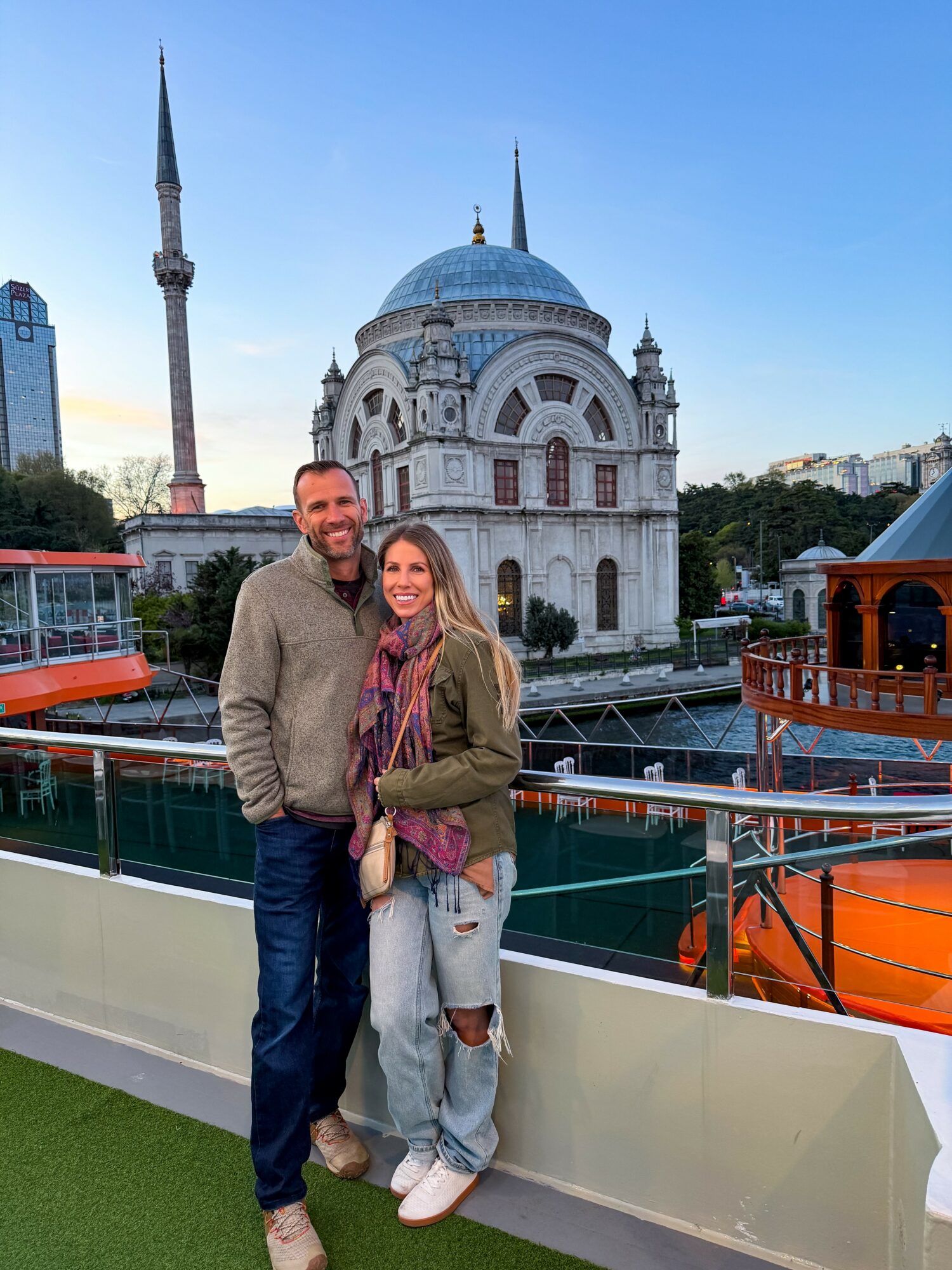 A man and woman stand together in front of a historic mosque with a large blue dome and minarets, on a boat deck.