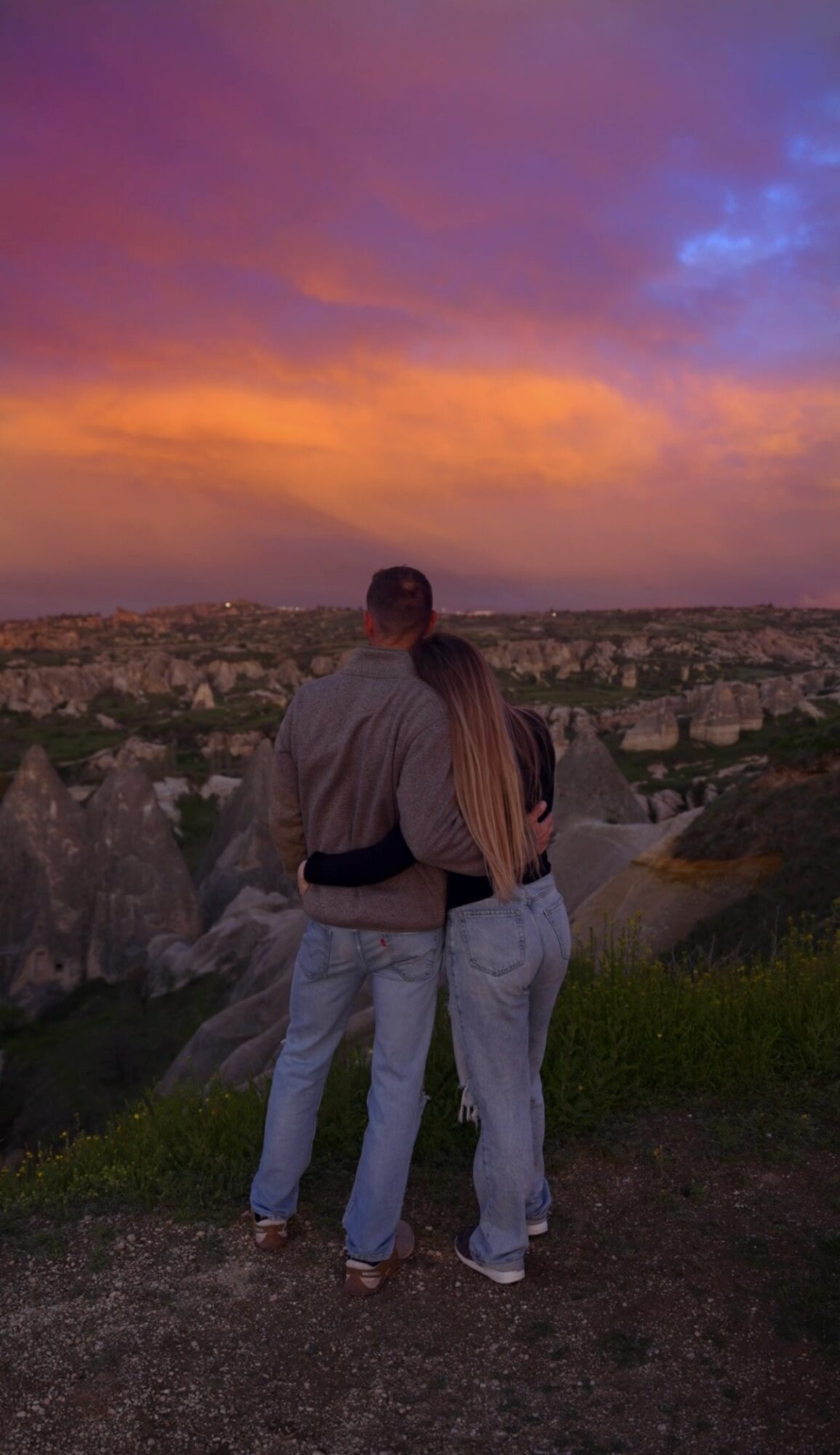 A couple stands on a grassy hill overlooking a cityscape at sunset, with colorful sky and clouds.