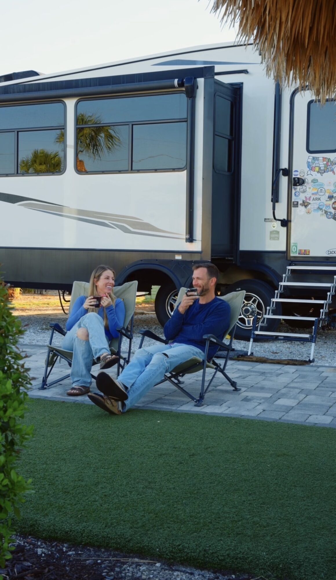 Two people sit outside near a large RV, talking and smiling, with a grassy area in the foreground.