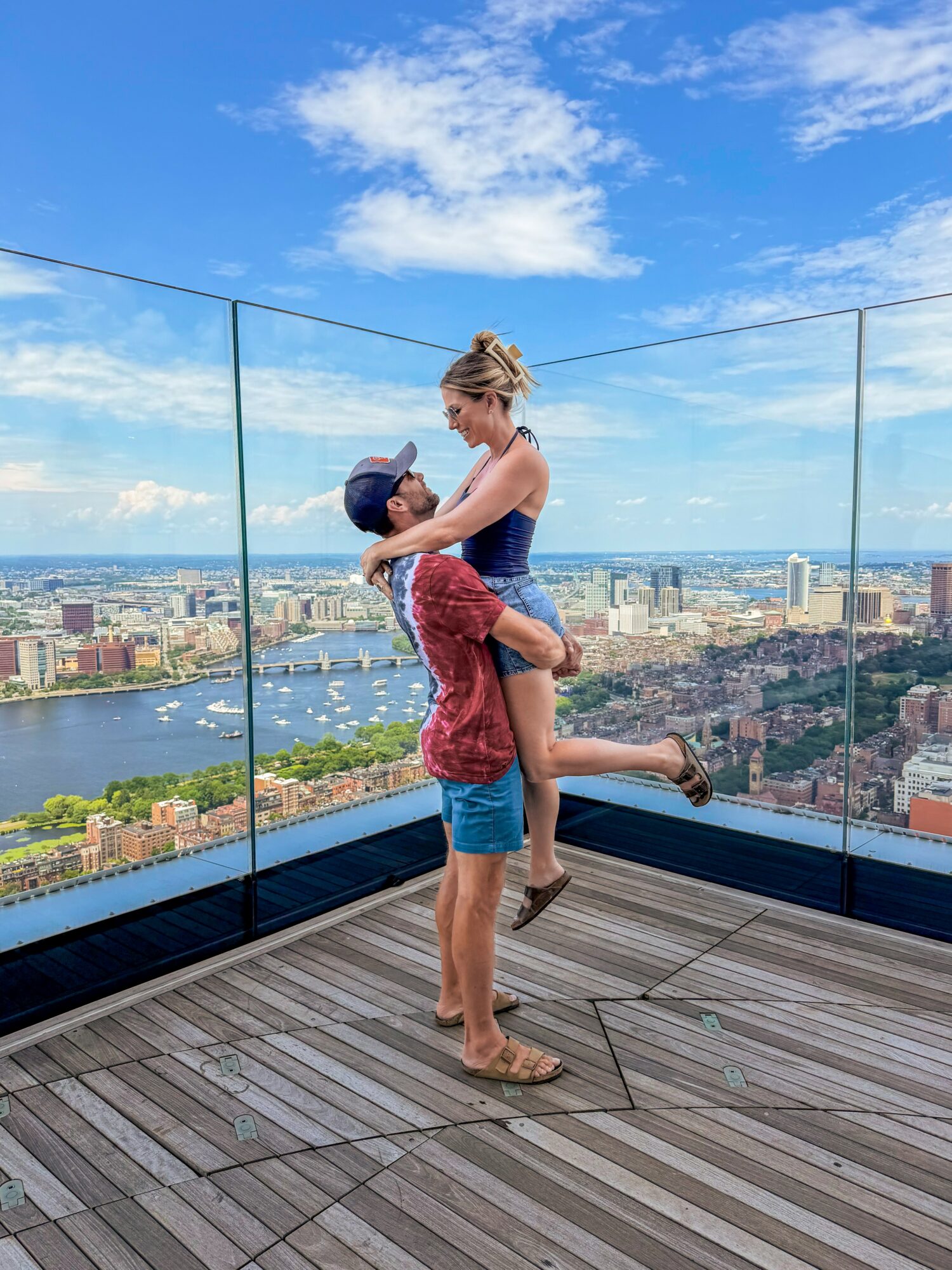 Two people on a rooftop with a city view, one lifting the other, both smiling, glass barriers behind them, wooden floor beneath.