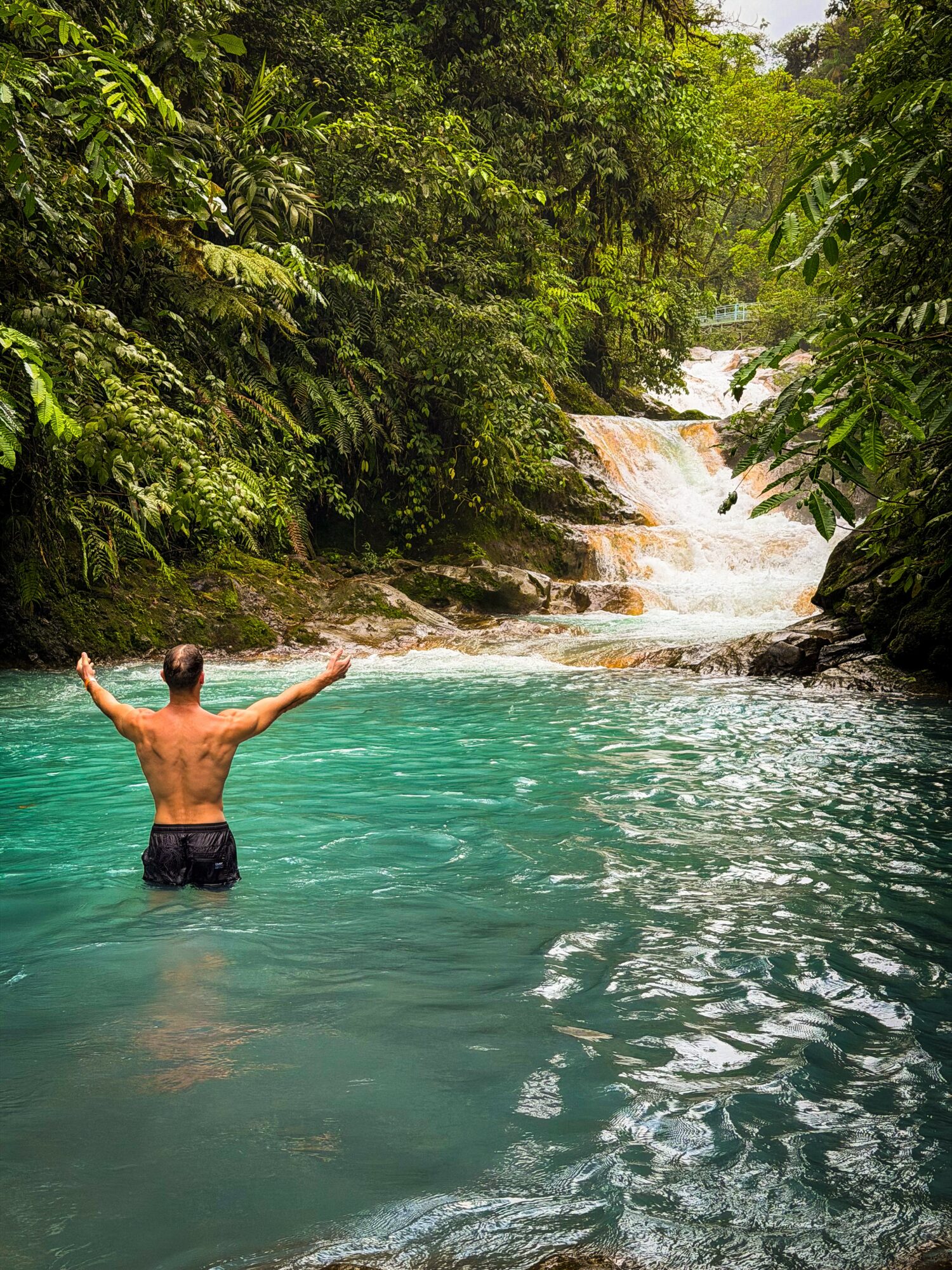 Person standing in turquoise water with arms raised, surrounded by lush green trees and a small waterfall in the background.
