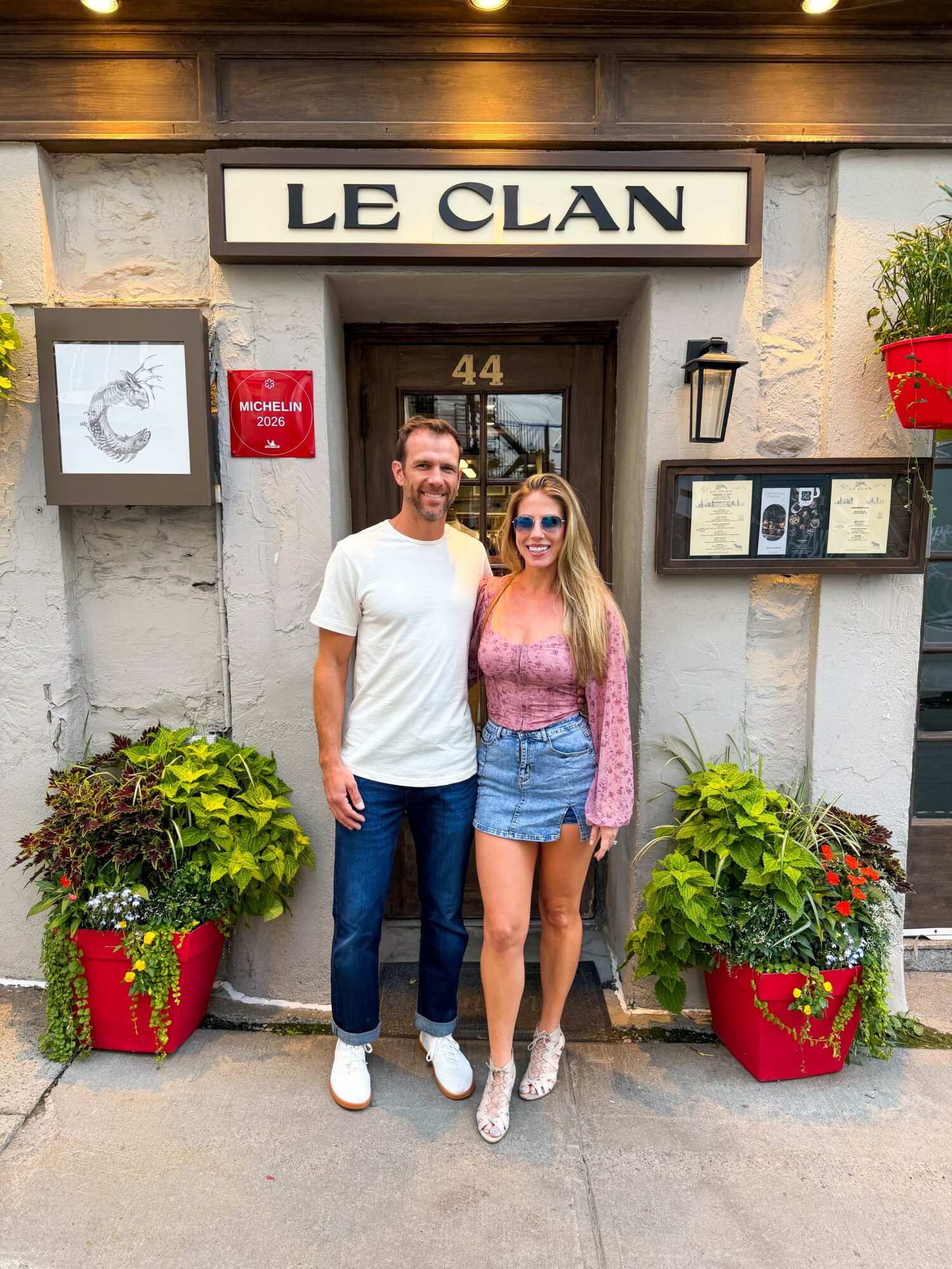 A man and woman stand in front of a building with a sign that reads 'LE CLAN,' surrounded by potted plants and decorative elements.