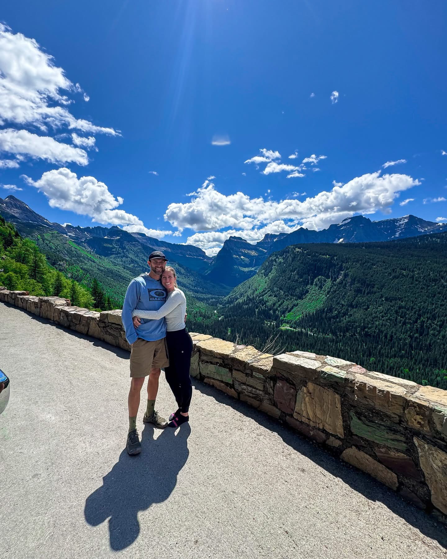 Two people standing on a mountain overlook with a valley and mountains in the background under a blue sky with clouds.