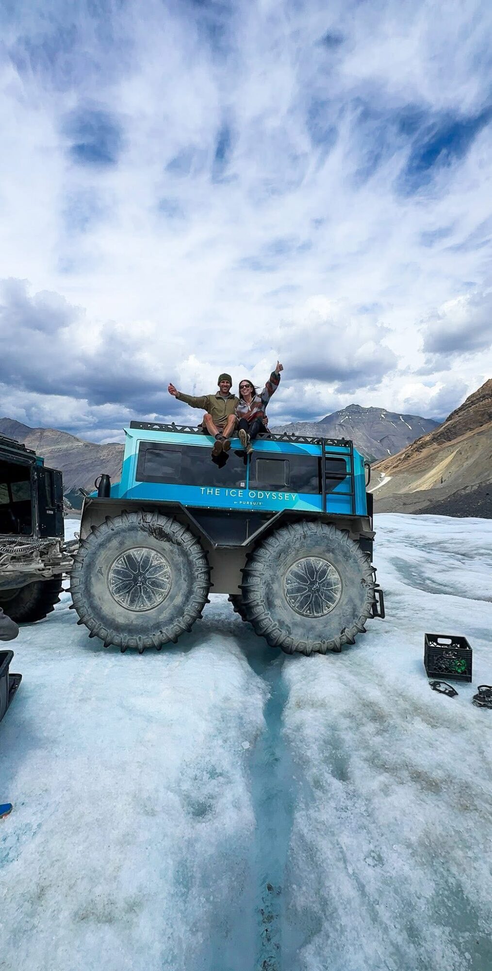 Person sitting on top of a blue vehicle in a snowy landscape with cloudy sky and mountains in the background.