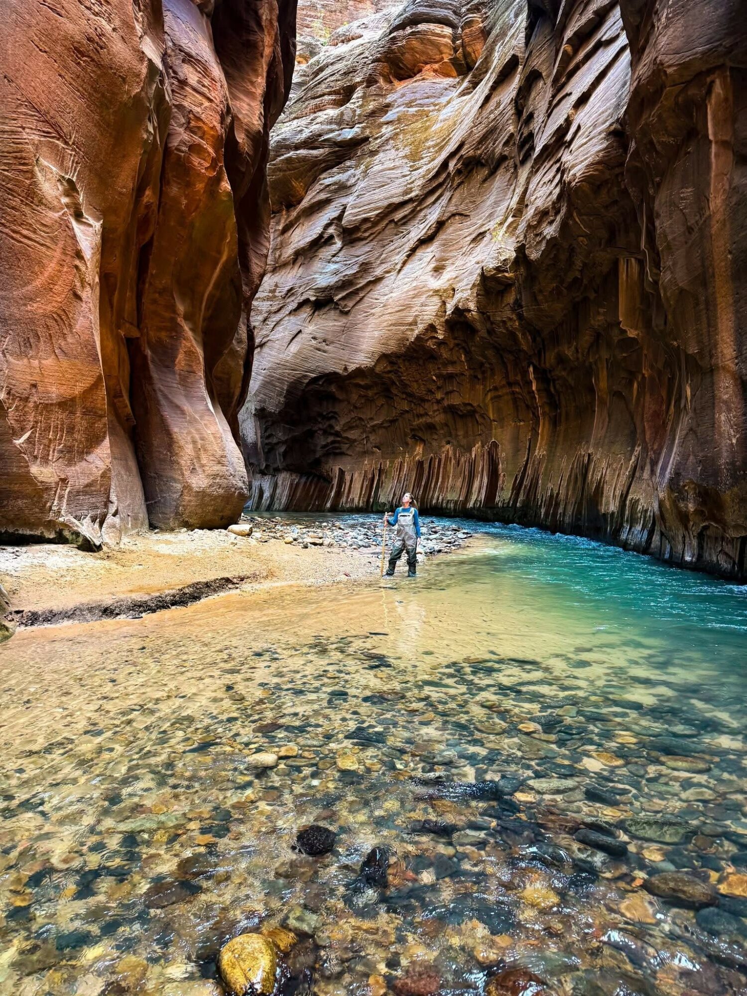 Person standing in a narrow canyon with colorful rock walls and a clear blue stream, sunlight filtering through.