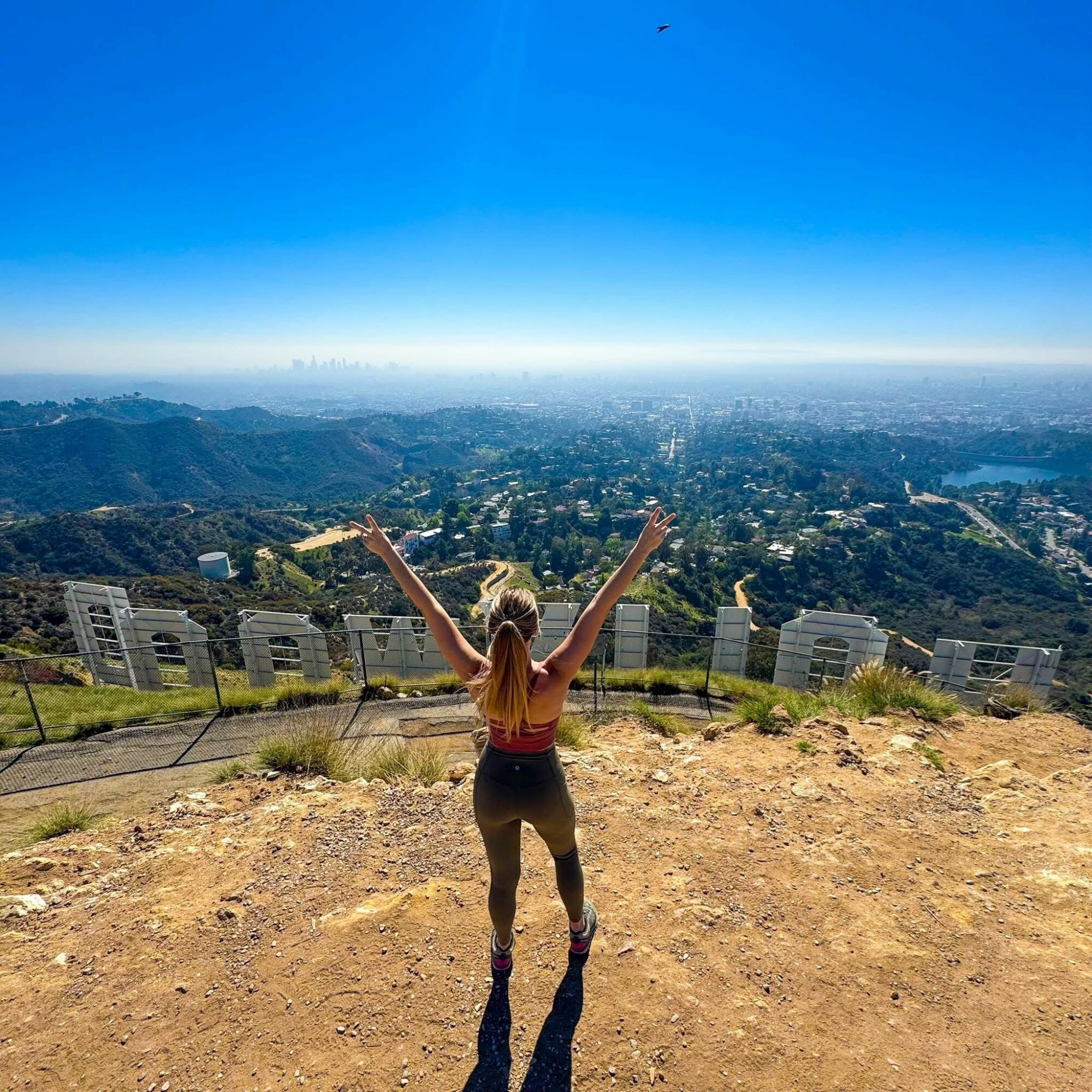 Person with long hair standing on a hilltop with arms raised, overlooking a cityscape and blue sky.