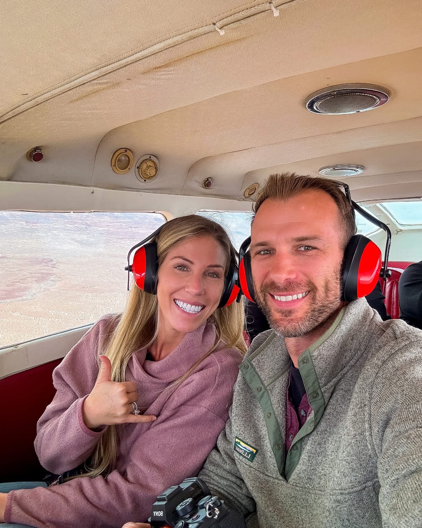 Two people smiling inside a helicopter, wearing headsets, with a landscape visible through the window.
