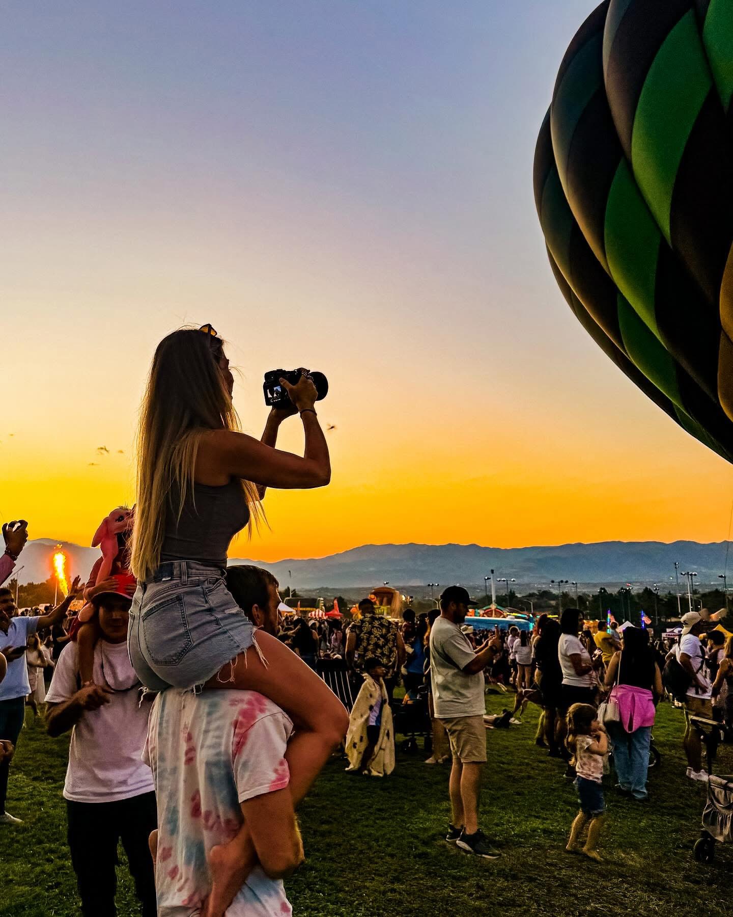 Girl sitting on a person's shoulders, taking a photo with a camera during sunset at an outdoor event with many people and a hot air balloon.