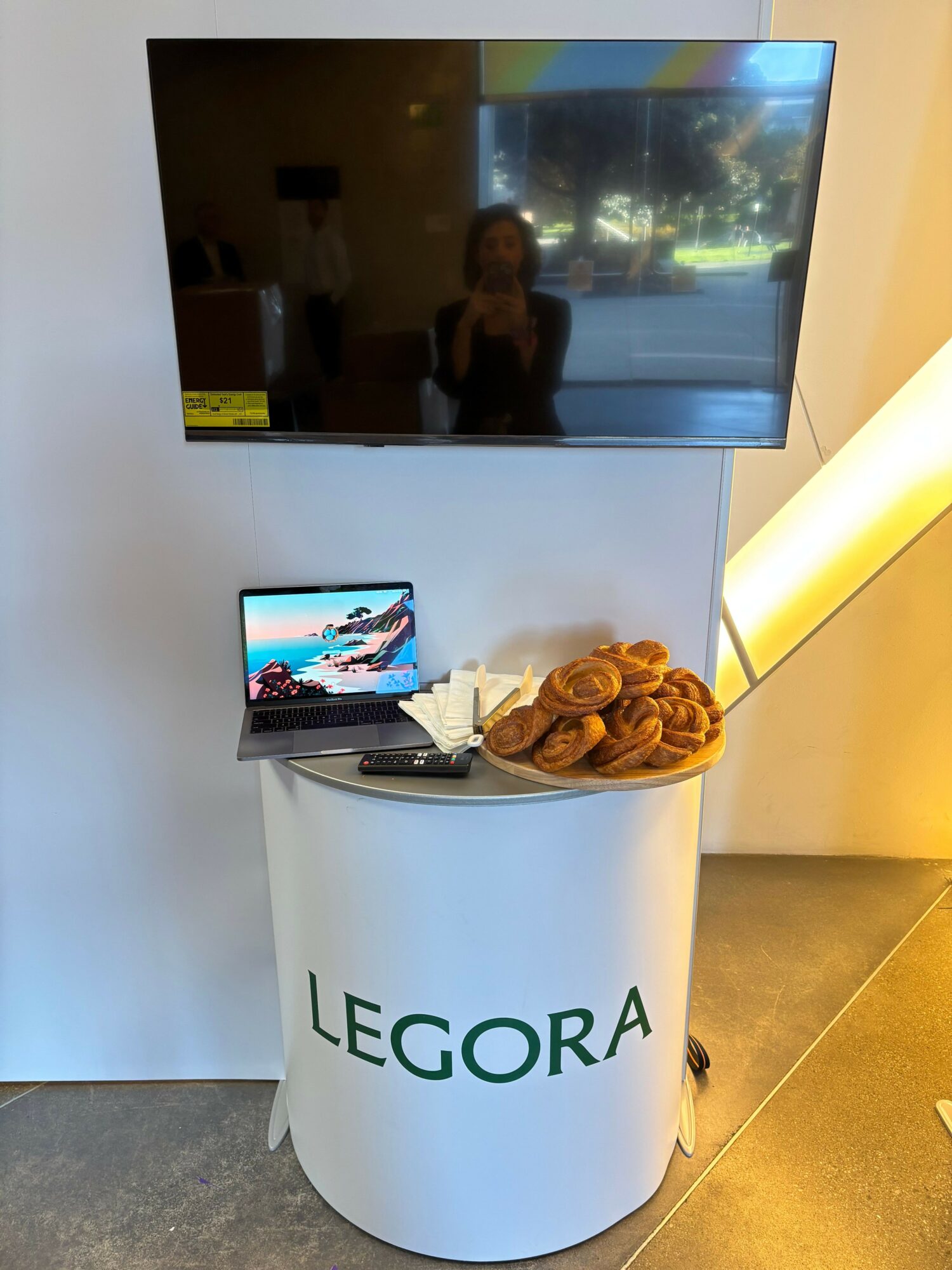 A white cylindrical display with the word 'LEGORA' and a plate of pastries, a tablet, and a TV screen.