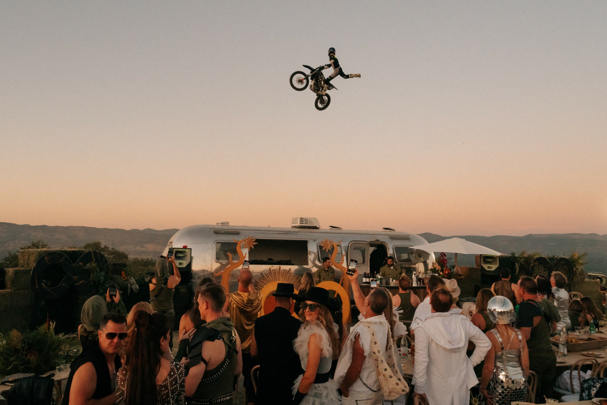 BMX rider mid-air above a crowd at an outdoor event during sunset, with tents and people watching.