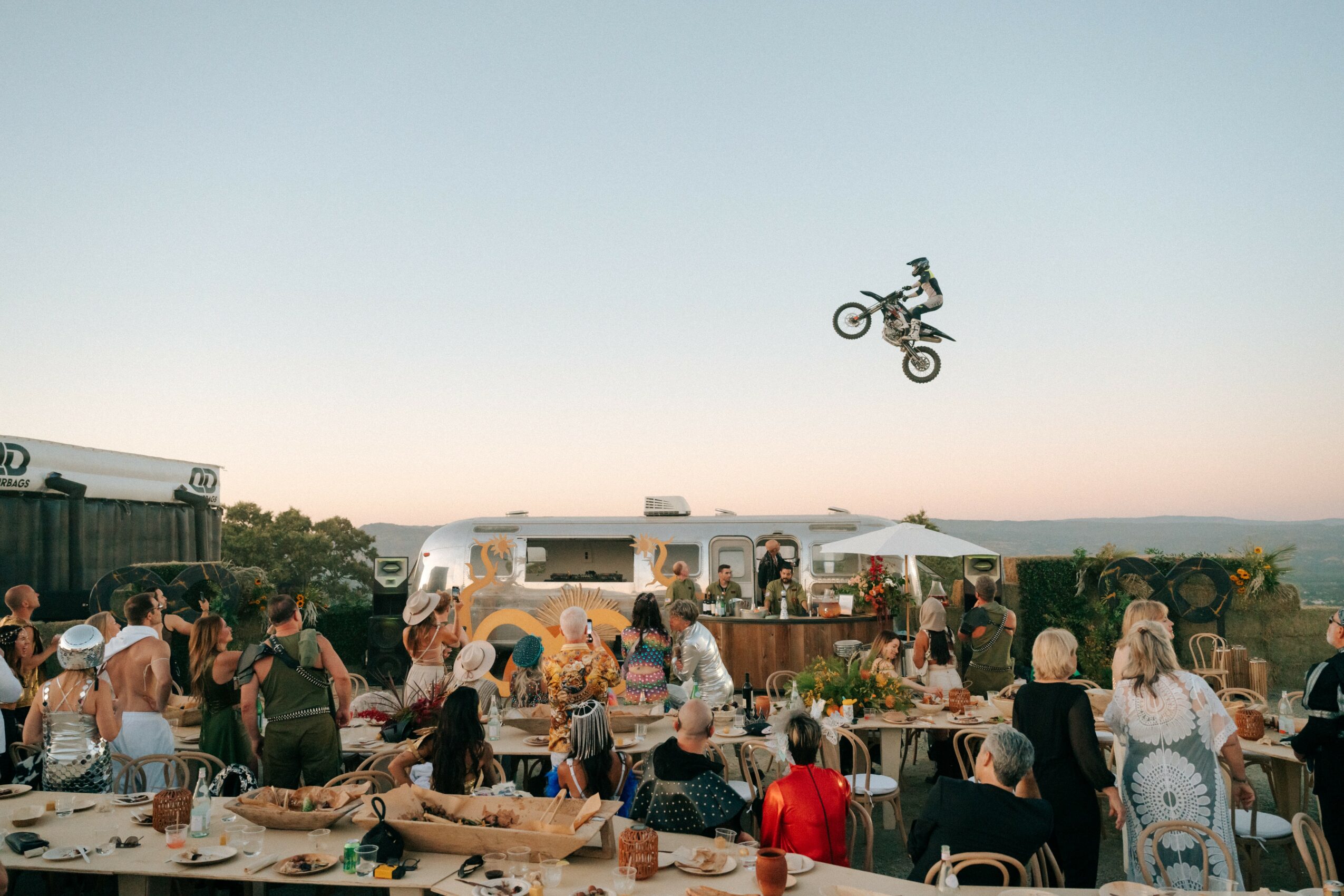 People gathered outdoors with a person performing a stunt on a bicycle in the sky, tents and a scenic background visible.