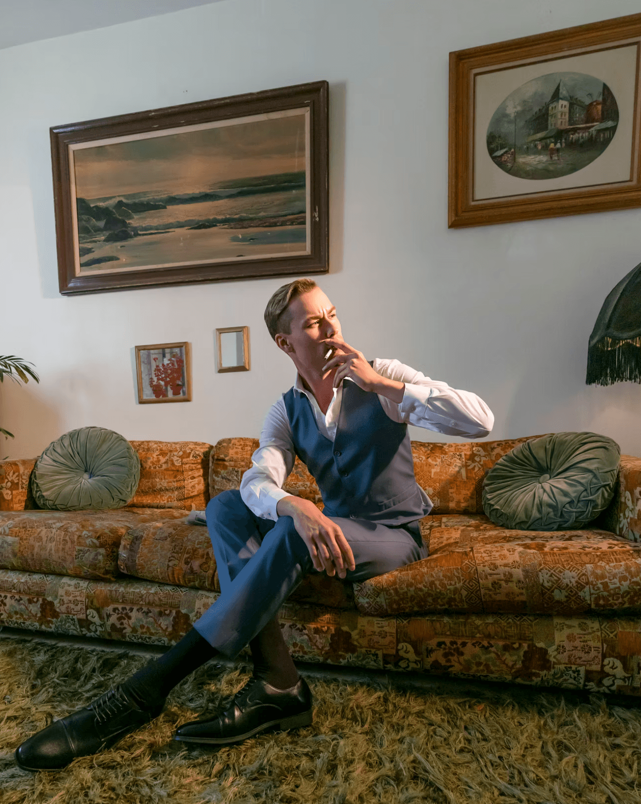 Man sitting on a patterned sofa with green pillows, in a room with framed paintings on the wall, resting his chin on his hand.